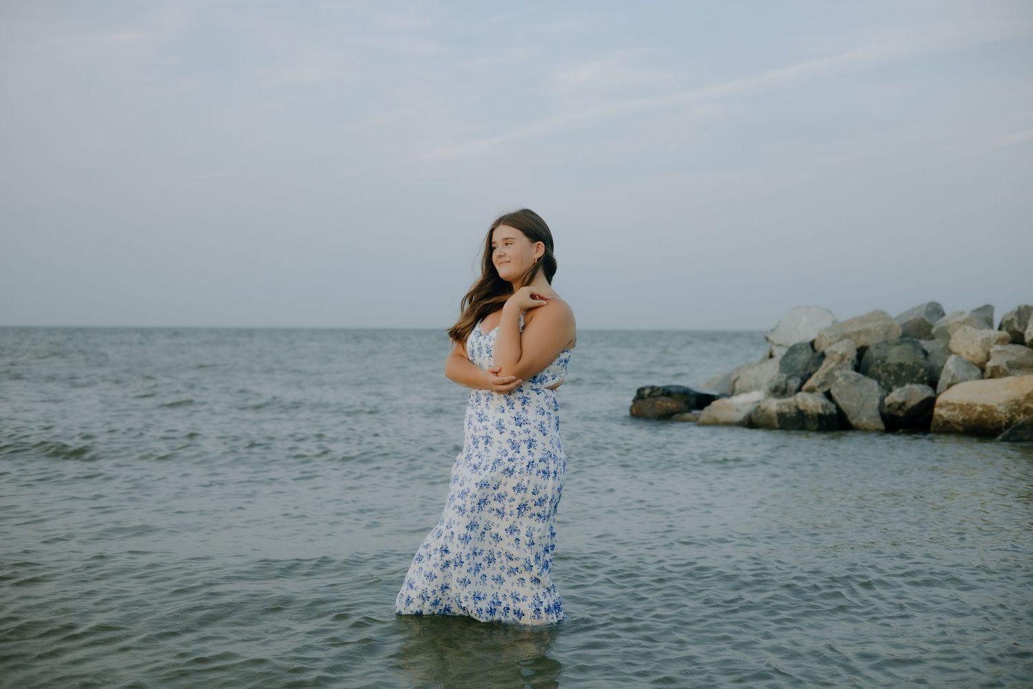 A person in a floral dress poses by the ocean with rocky outcrops visible in the background.