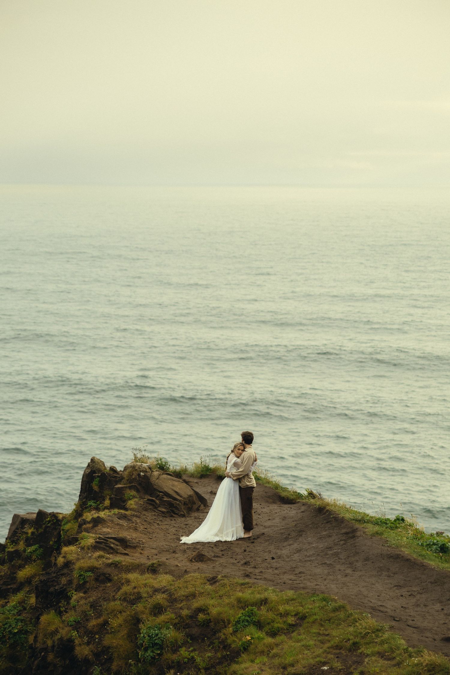 Bride and groom embracing on a rocky coastal cliff overlooking a calm misty ocean on their wedding day.