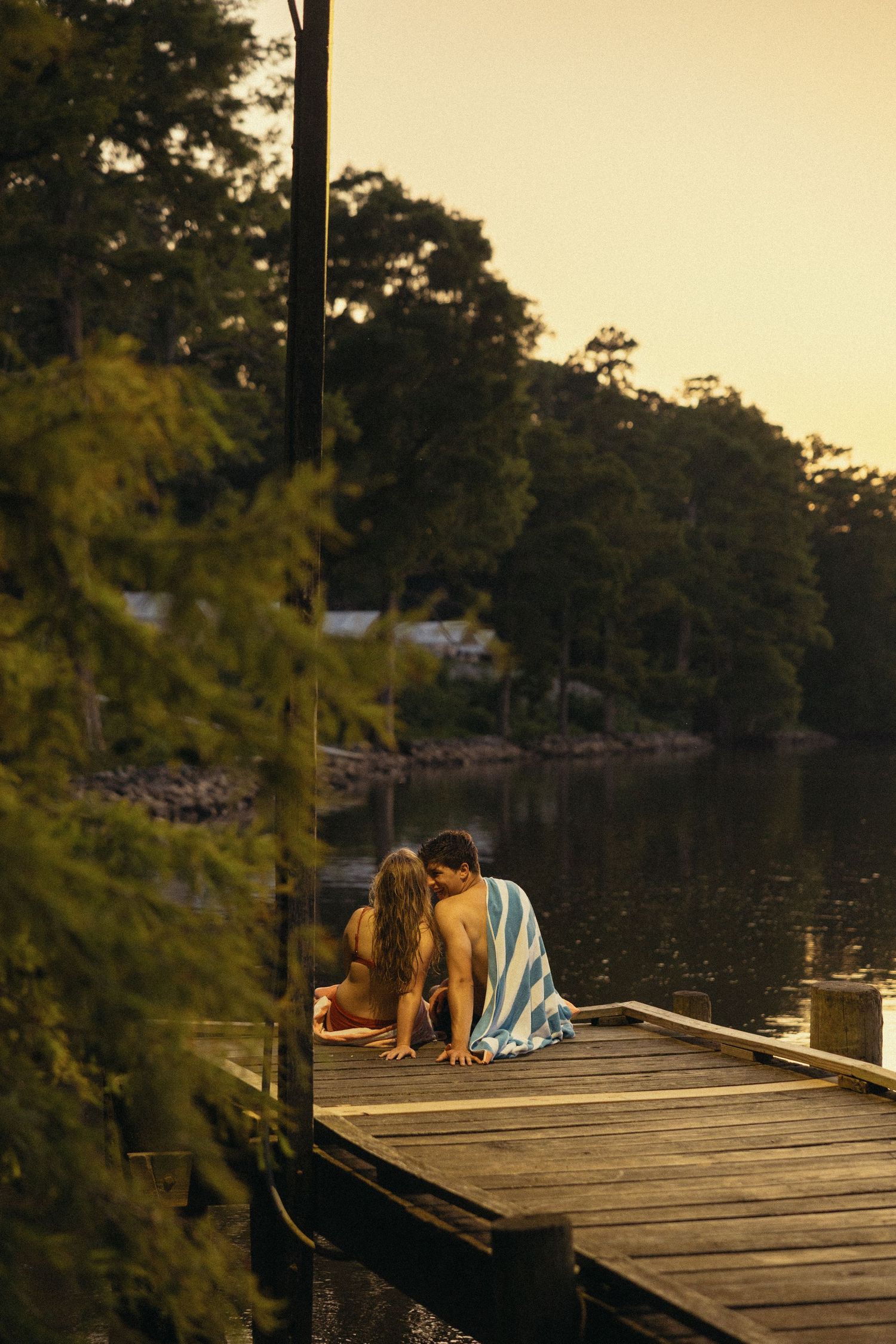 Couple sharing a romantic kiss on a wooden dock by a serene lake at sunset, wrapped in a striped towel.