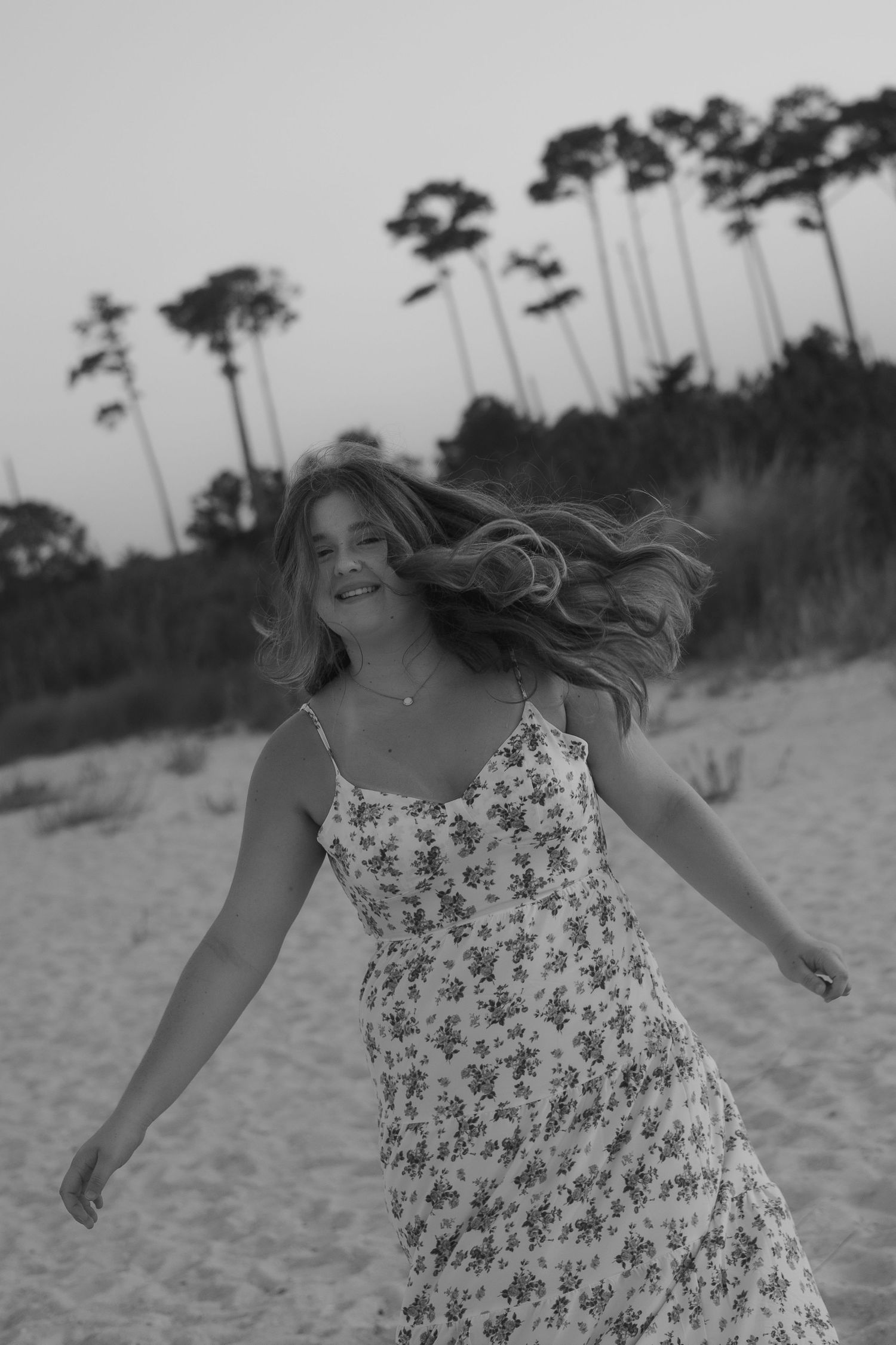 A person in a floral sundress runs joyfully along a sandy beach with tall pine trees in the background.