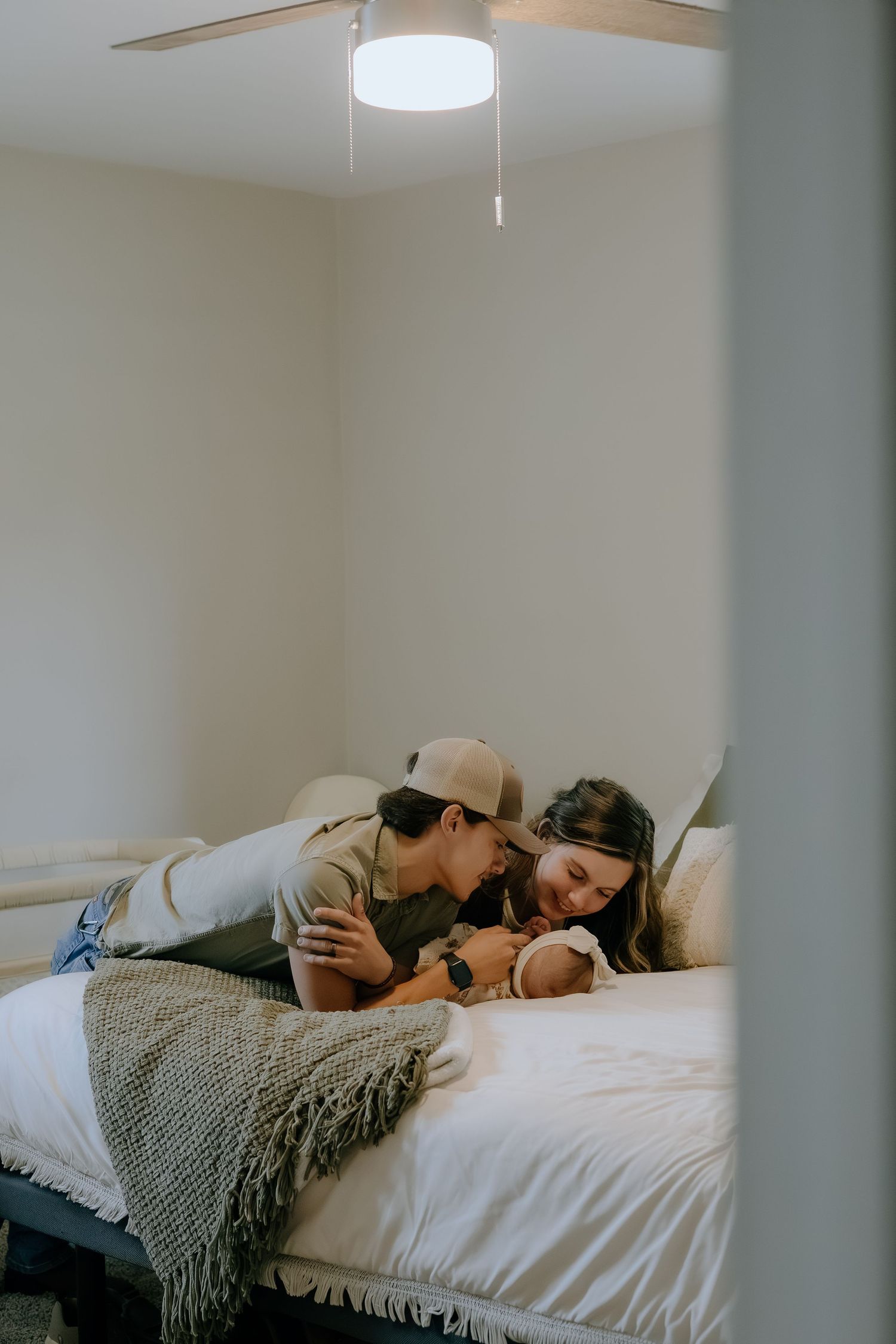 A couple rests together on a bed covered with a gray blanket in a bright room.