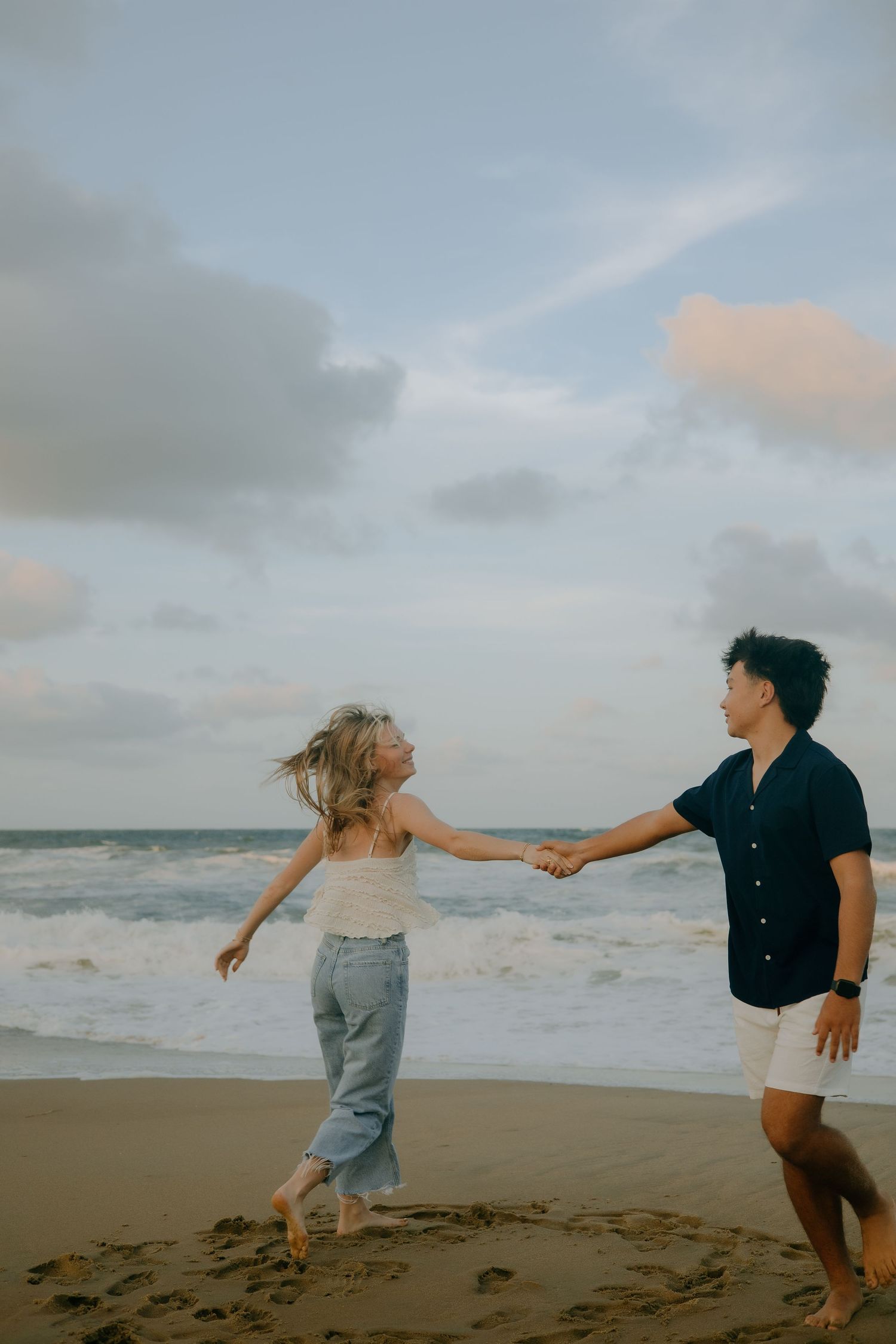 A couple holds hands while playfully dancing on a misty beach at sunset.
