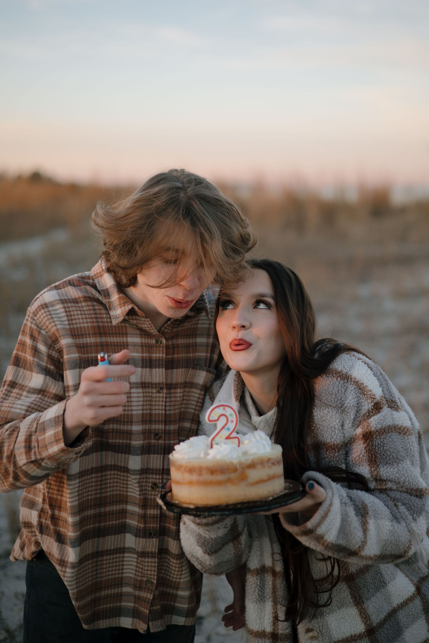 A couple in cozy winter clothing share a birthday donut with a candle during sunset outdoors.