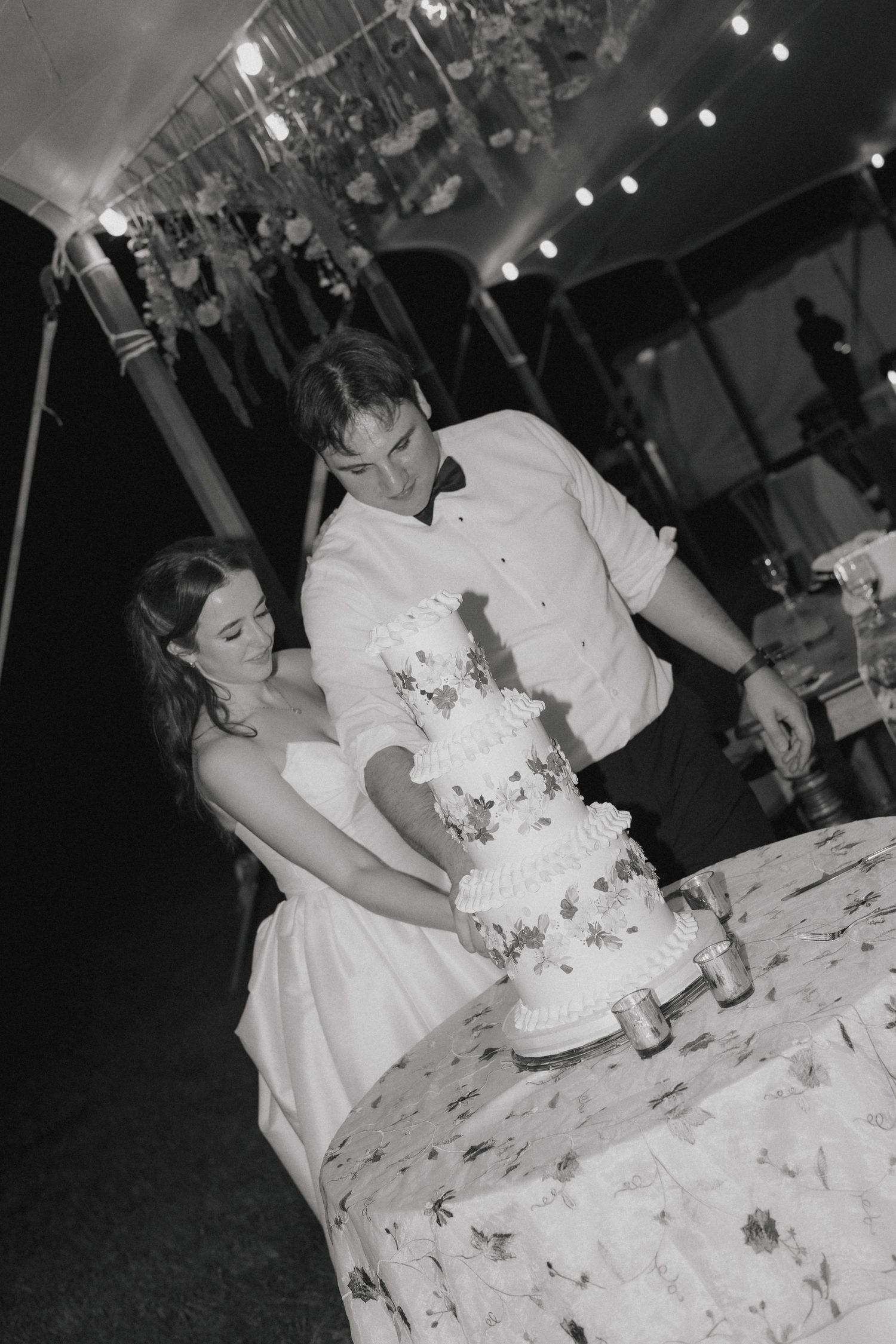 A black and white photo captures newlyweds cutting their elegant tiered wedding cake together.