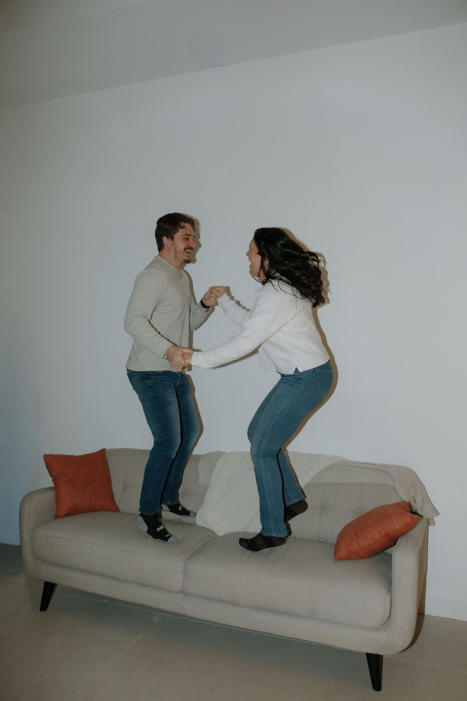 A joyful couple dancing together on a beige sofa in a modern living room with orange accent pillows.