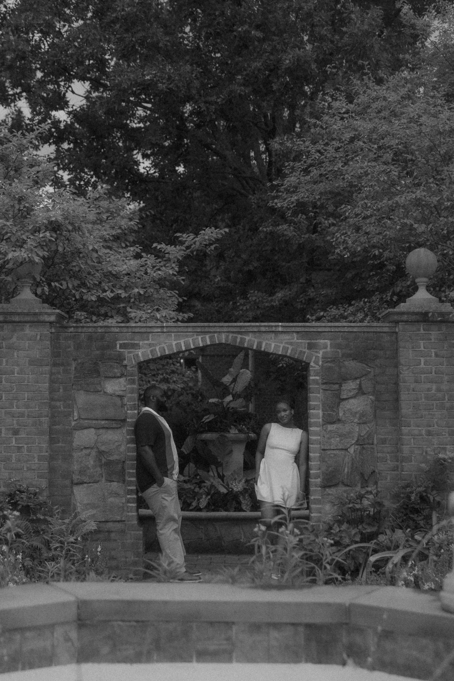 Black and white photo of a couple standing in a brick garden archway surrounded by lush trees and foliage.