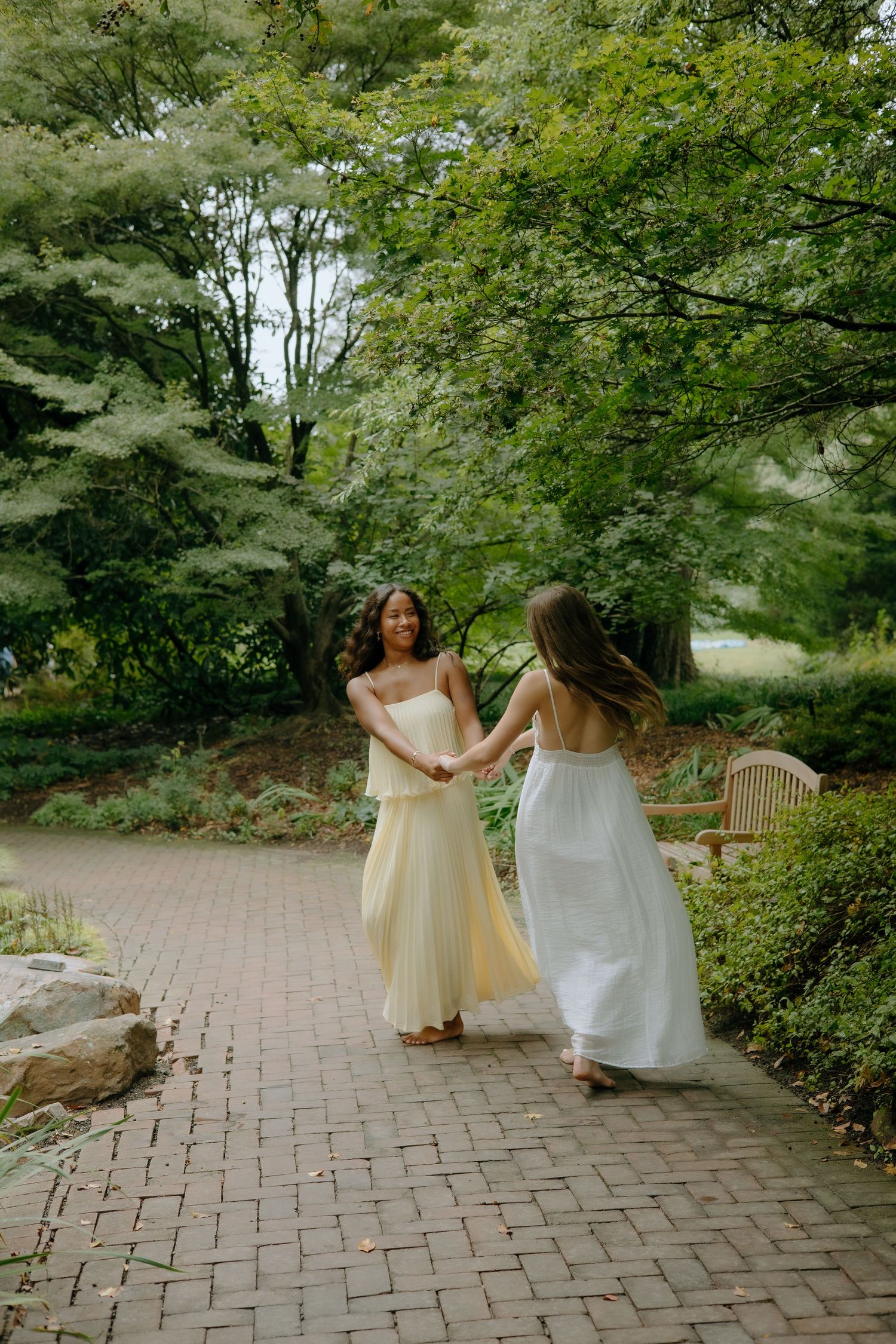 People in light-colored dresses twirl and dance along a brick pathway in a garden.