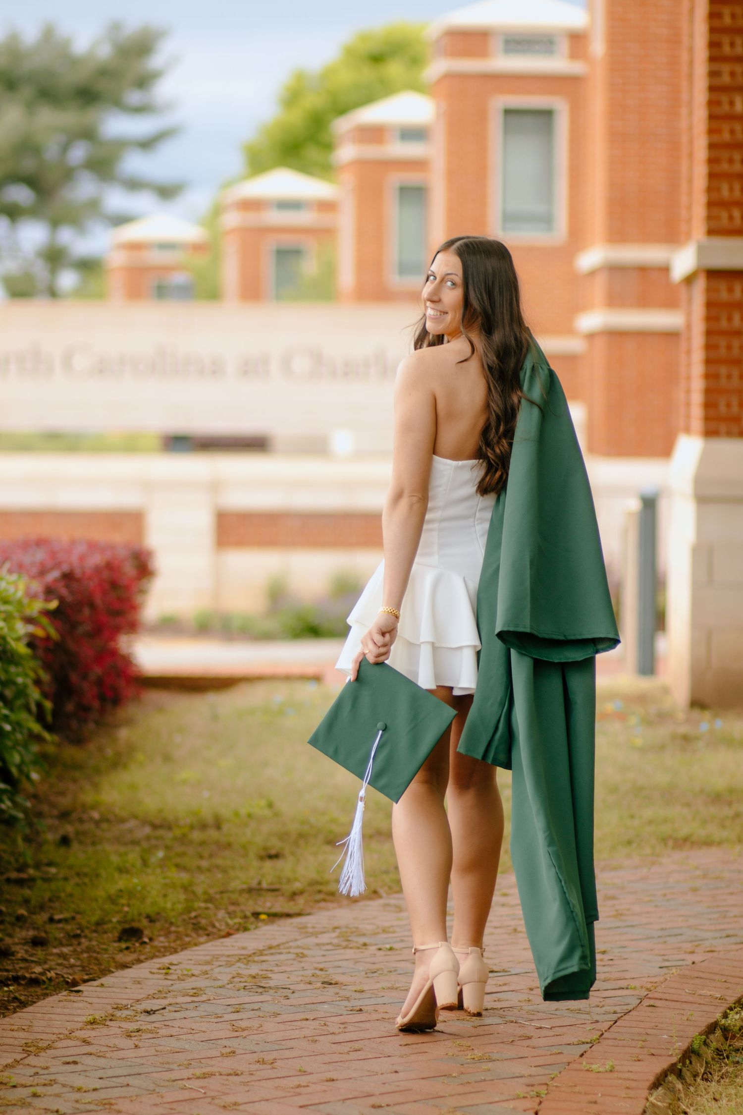 Graduate in white dress holds green cap and gown while standing on brick pathway.