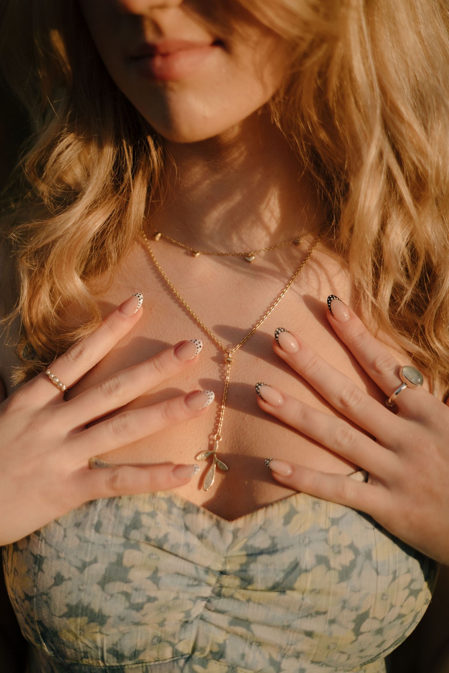 A person wearing a floral print dress touches their chest while wearing multiple rings and a delicate necklace.