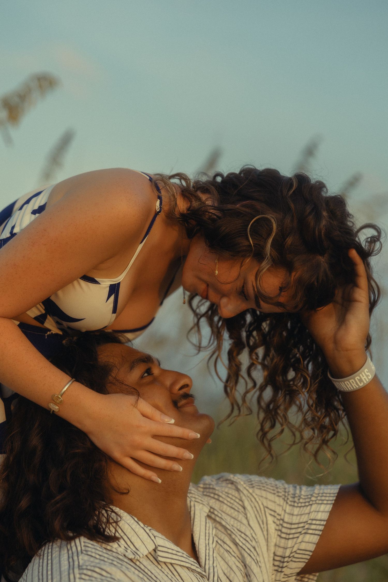 Two curly-haired women share an intimate moment outdoors in a field at golden hour, one leaning over the other smiling.