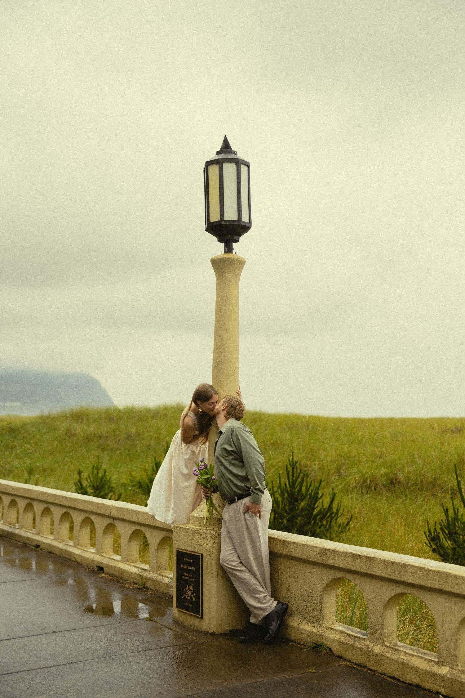 Couple sharing a romantic kiss by a lamppost on a rainy terrace overlooking green fields and ocean.