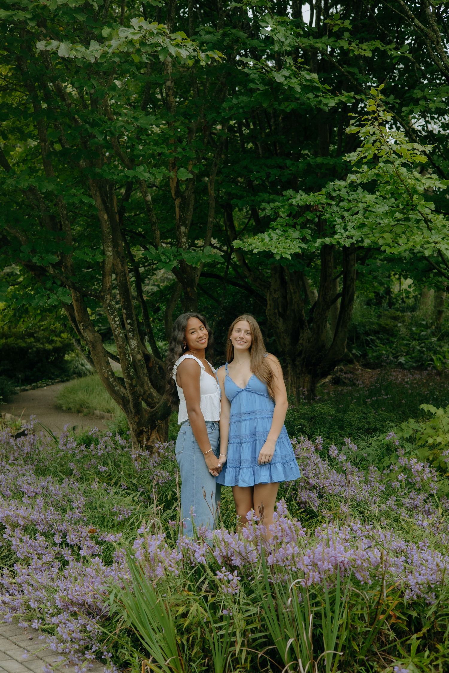 Two young women smile in a lush garden surrounded by purple flowers and green trees.