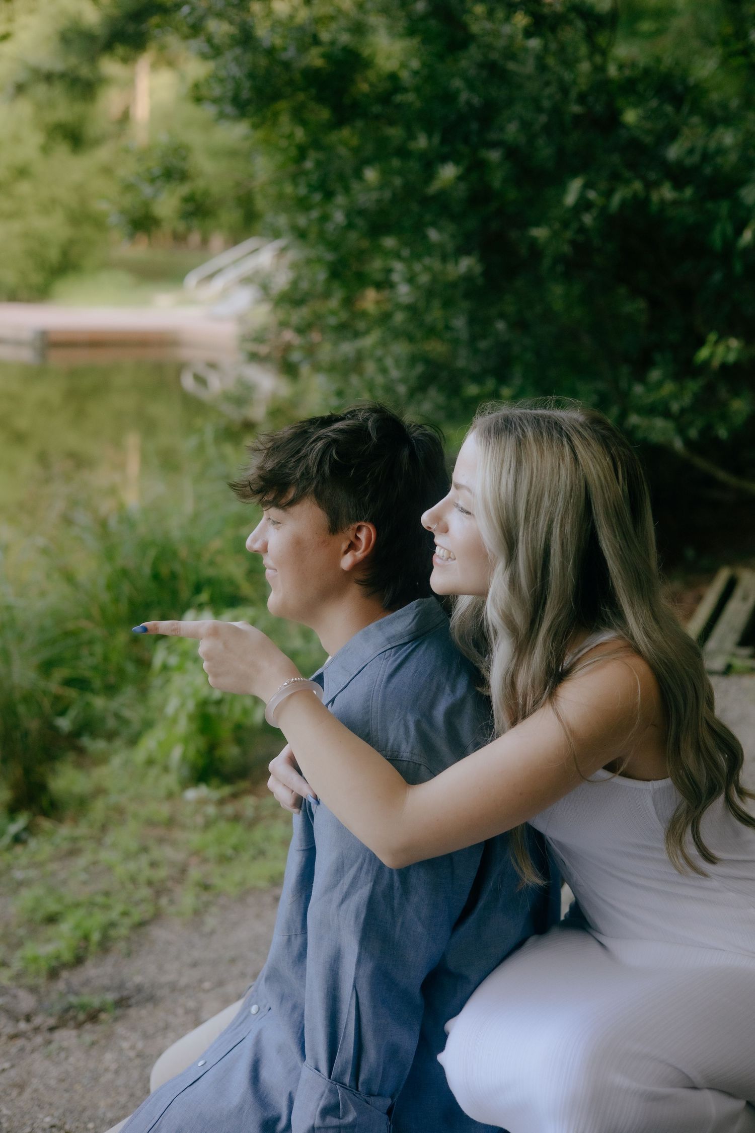 A young couple sits outdoors, the woman hugging the man from behind as she points toward a lush green landscape.