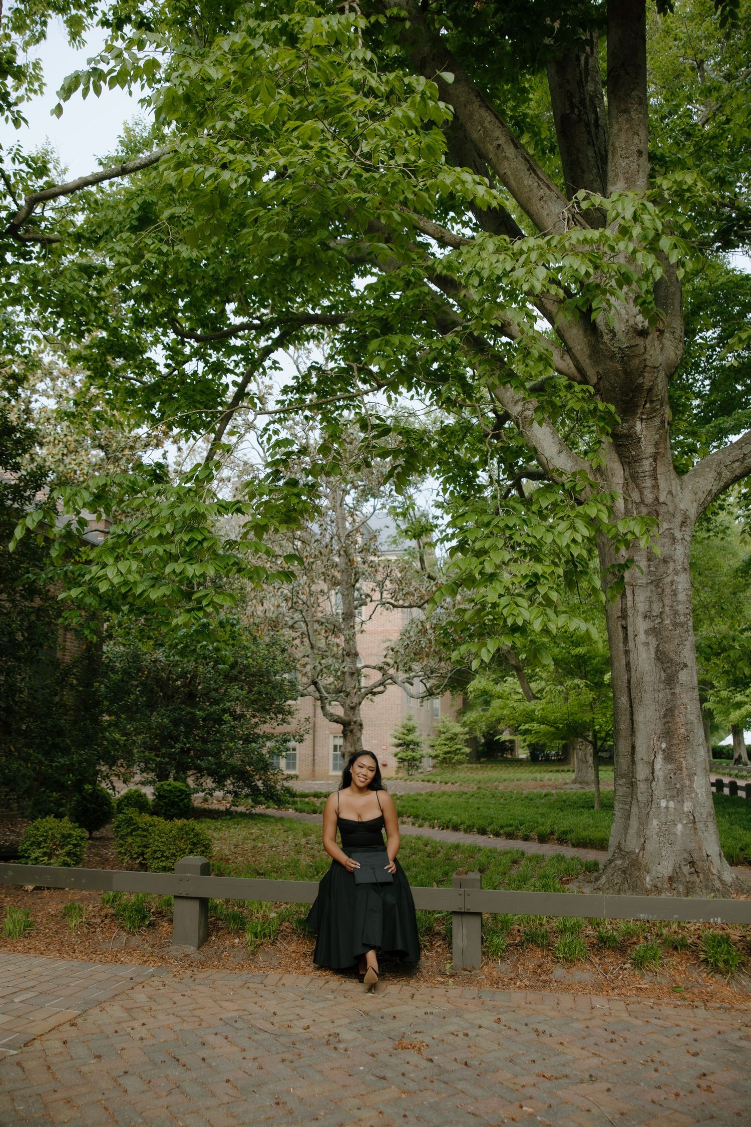 Woman in black gown sitting on bench under large green tree on brick path near historic building.