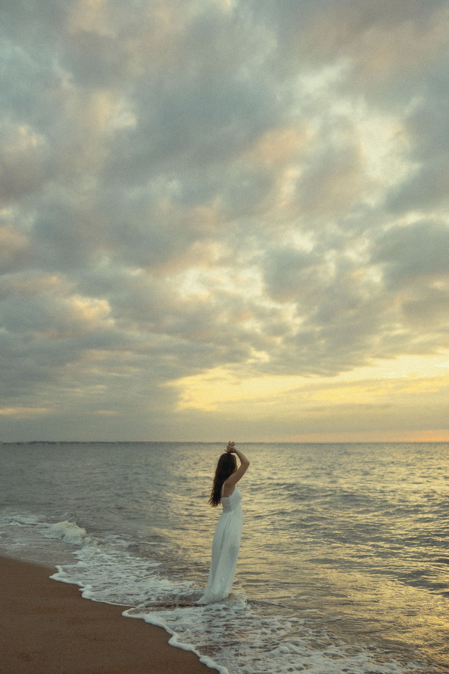 Woman in white dress standing at ocean shore during golden sunset with dramatic cloudy sky.