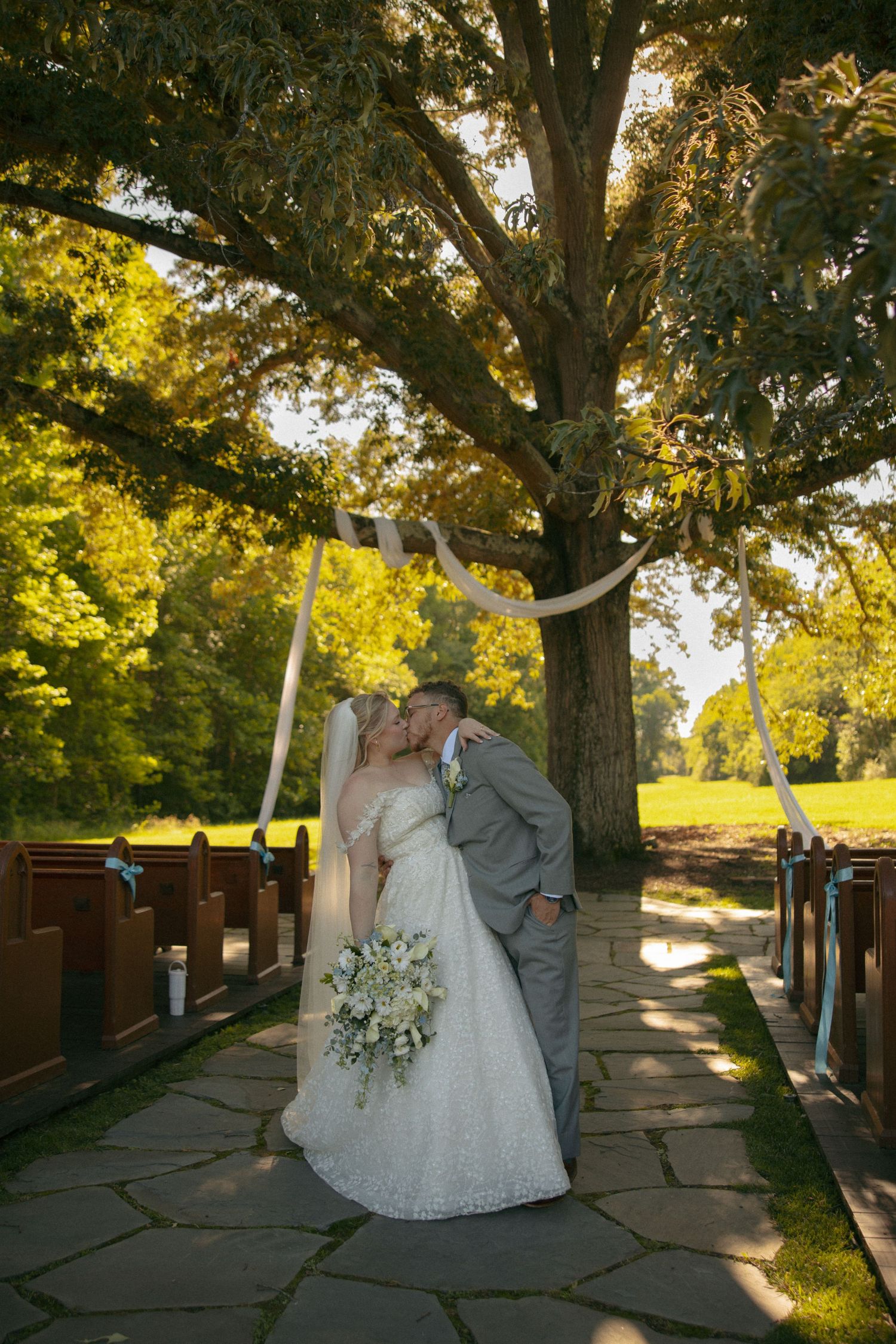 Bride and groom share first kiss under decorated tree arch on stone pathway at outdoor wedding ceremony.