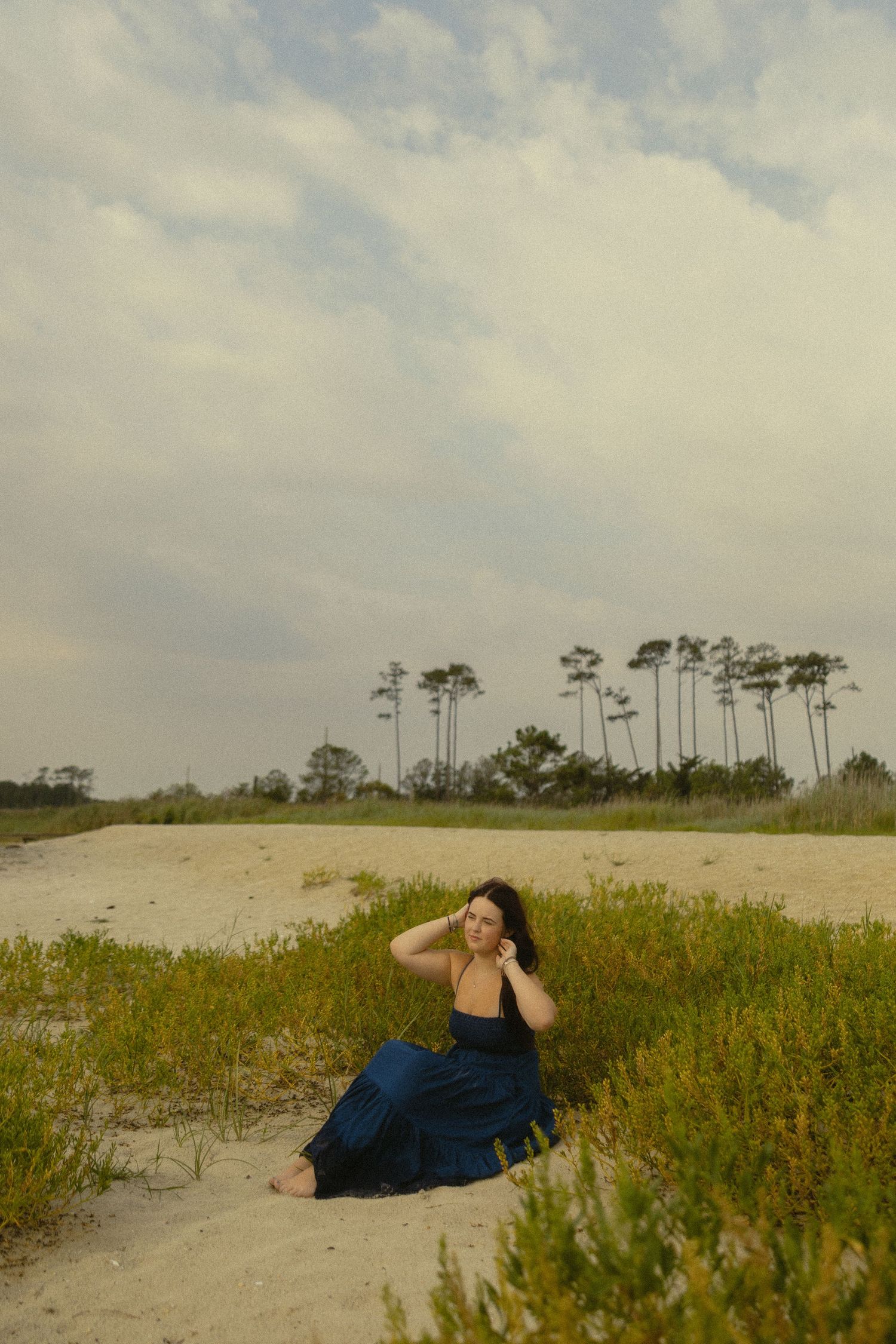 Woman in blue dress sitting on sandy beach dunes with tall pine trees and cloudy sky in background.