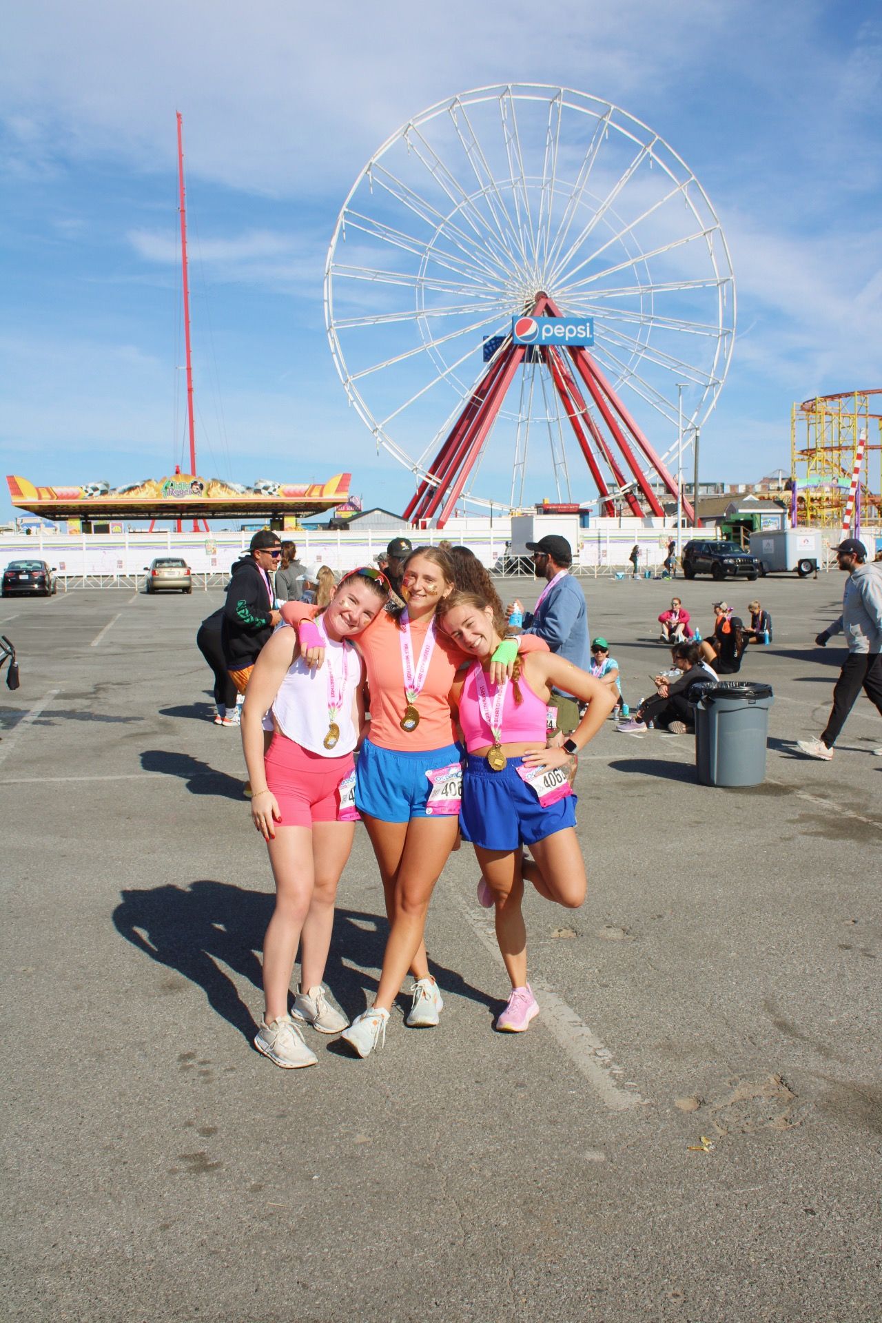 Three girls in pink athletic wear pose with medals near a ferris wheel at an amusement park after a fun run race.