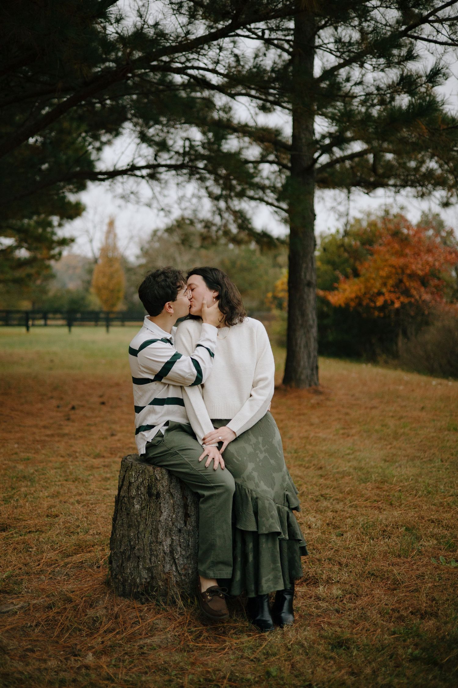 A couple shares an intimate moment while sitting on a tree stump in an autumn park setting.