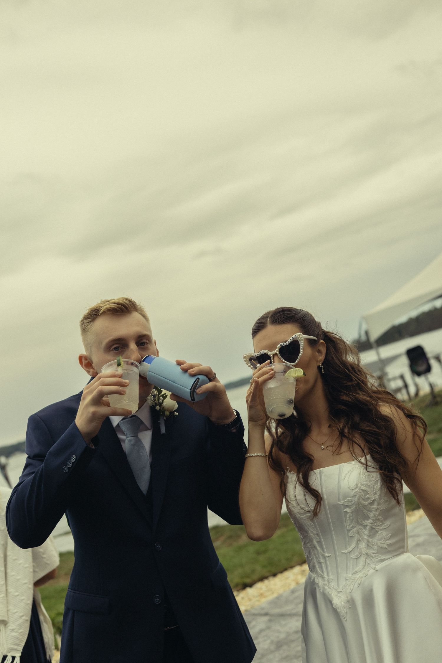 Bride in white dress and groom in navy suit drinking from cups at outdoor wedding celebration.