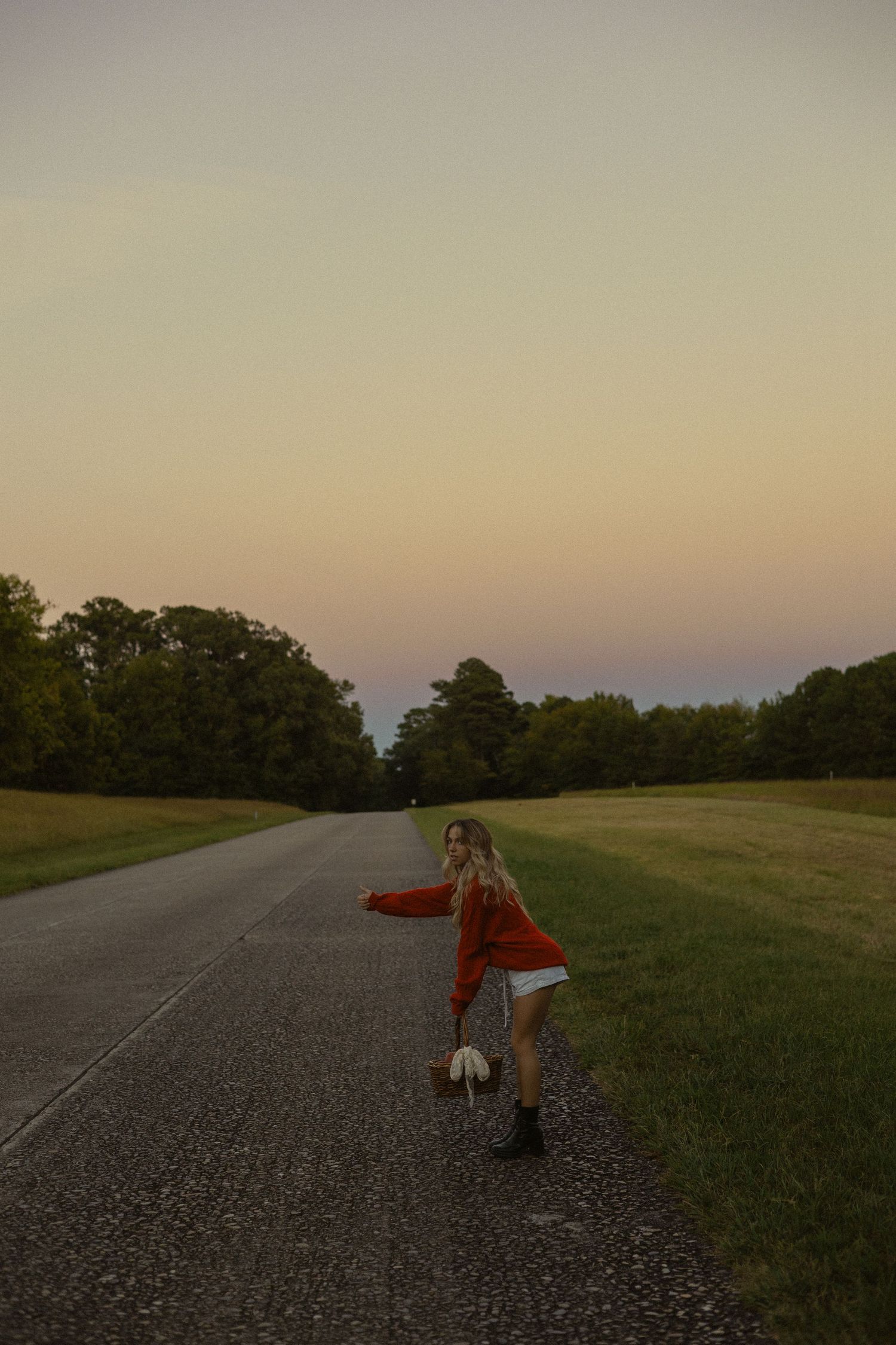 Woman in red top and white shorts hitchhiking on a rural road at sunset with trees in background.