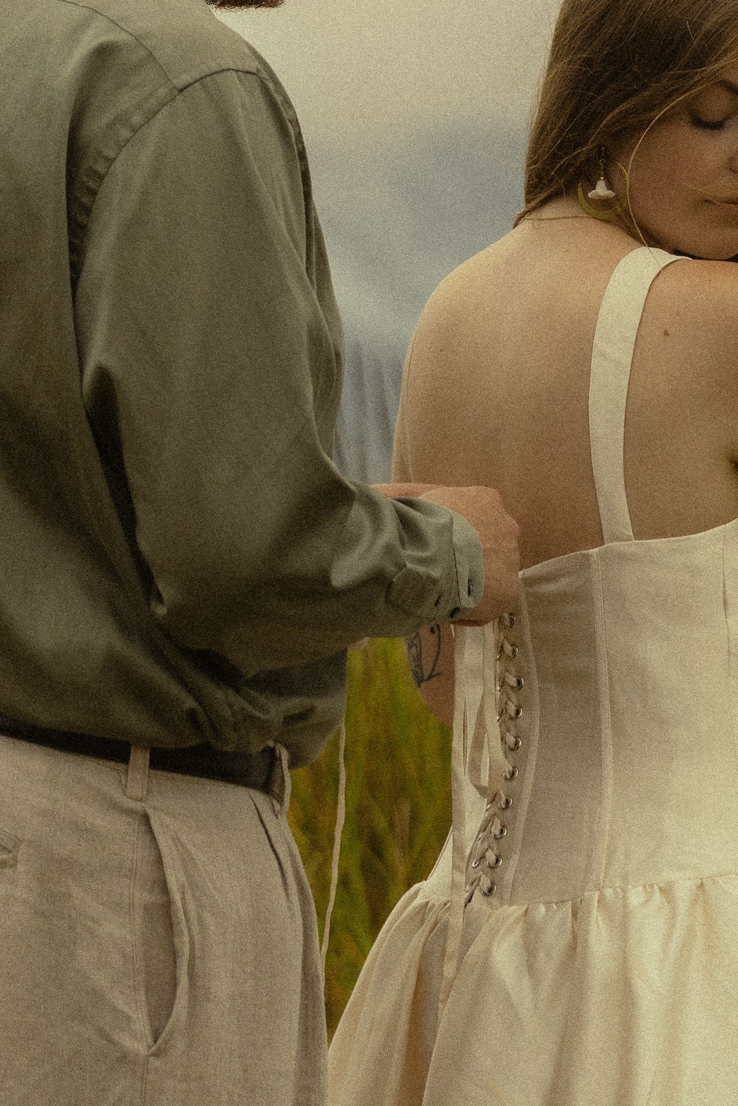 A man in olive shirt laces up the back of a woman's white dress outdoors.