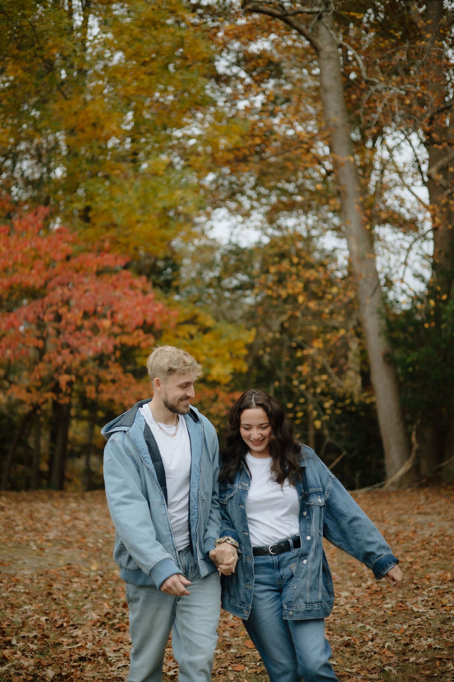 A couple shares a playful moment while walking through a forest filled with colorful fall foliage.