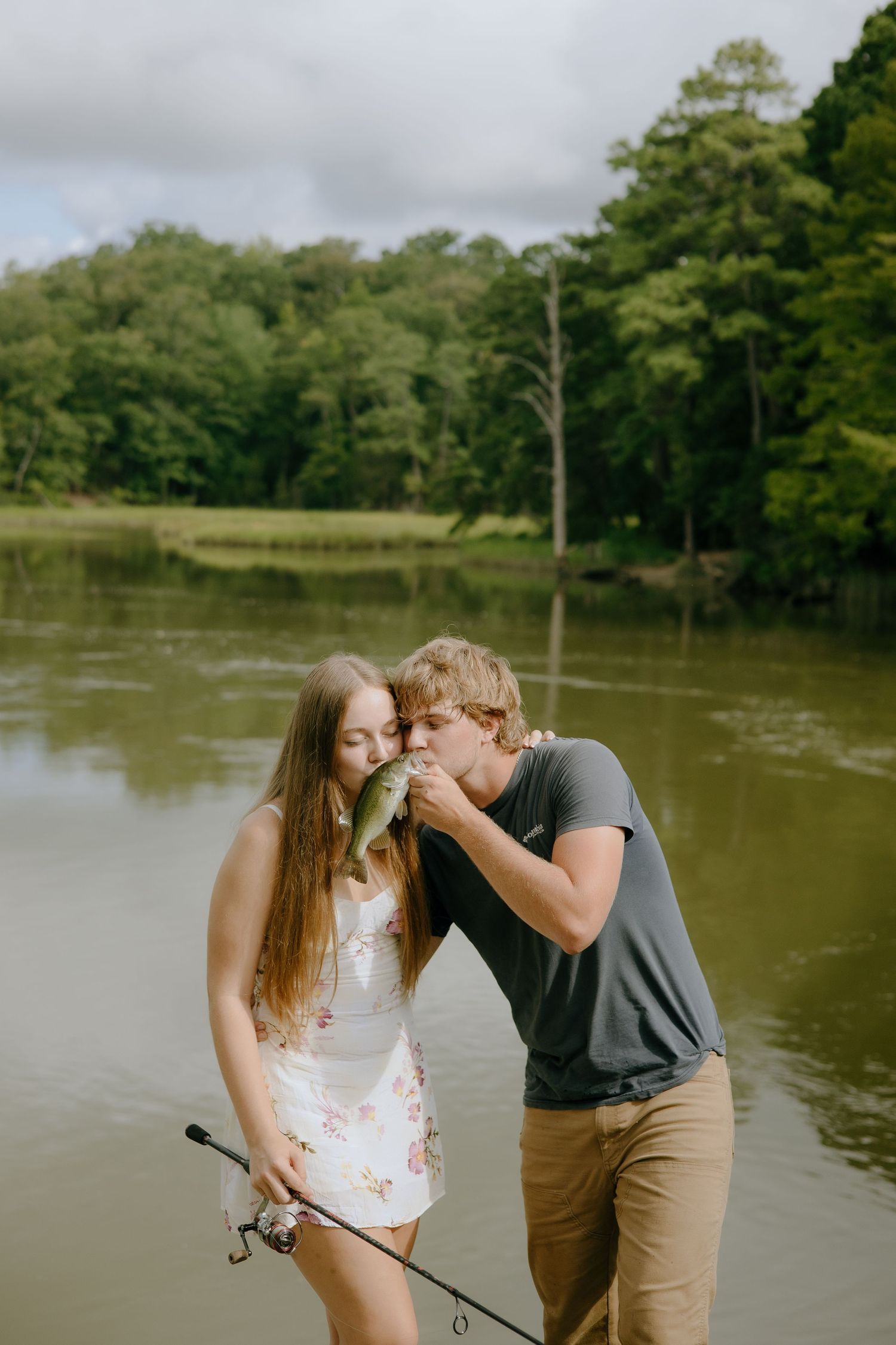 A romantic moment unfolds between a couple while fishing by a river surrounded by trees.