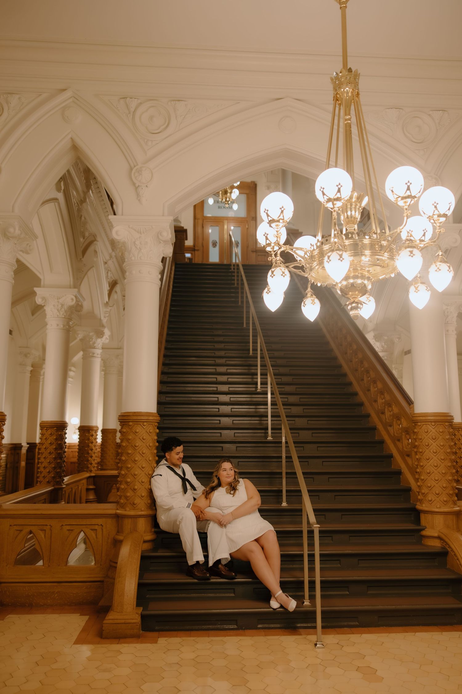 Couple in white outfits sitting on grand staircase in elegant venue with ornate chandelier.