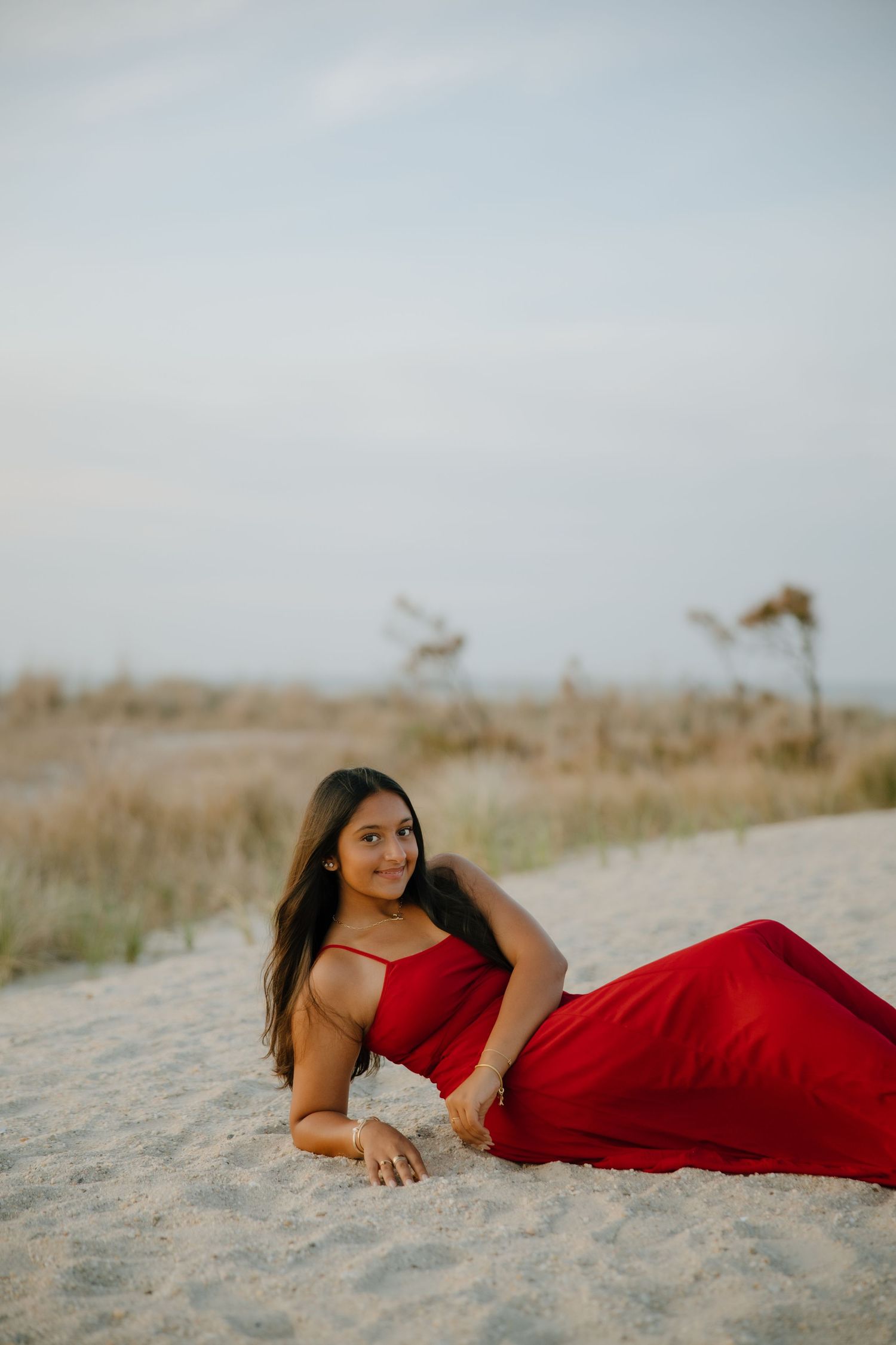 Young woman in red dress posing on sandy beach dunes with coastal grasses in background.
