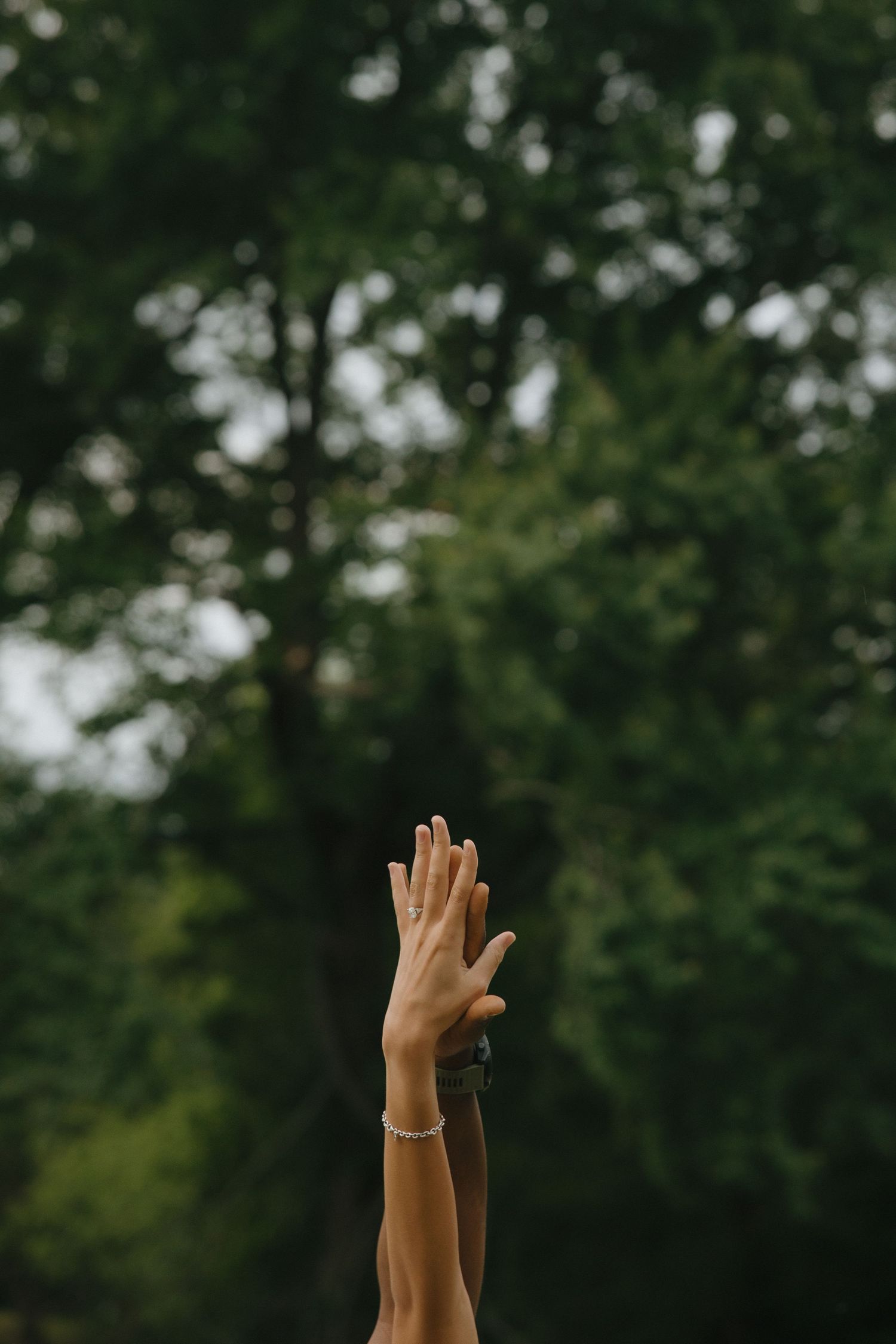 Two hands raised up against a lush green forest background, one wearing a delicate pearl bracelet.