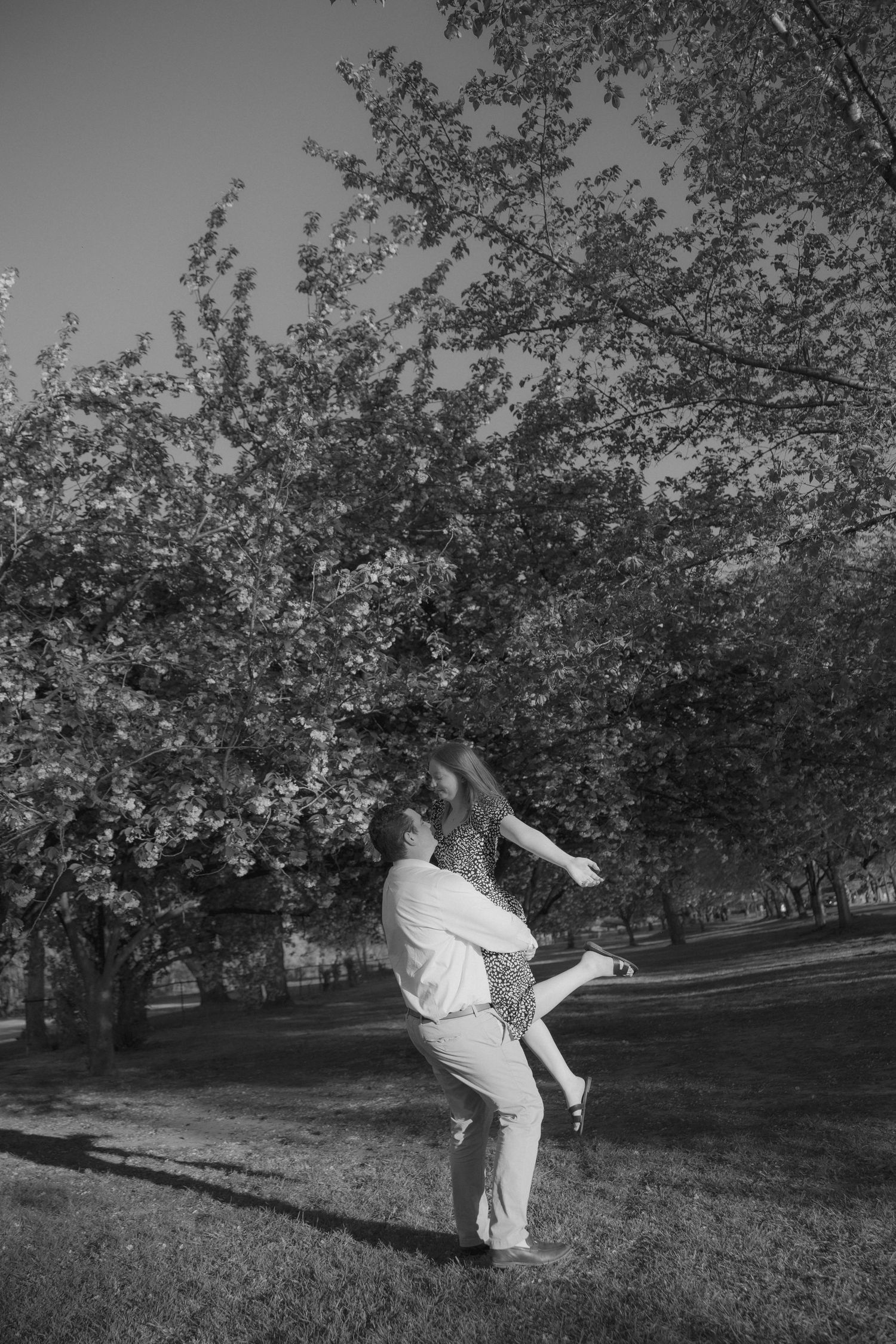 Black and white photo of a couple dancing joyfully in a park under blooming trees on a sunny day.
