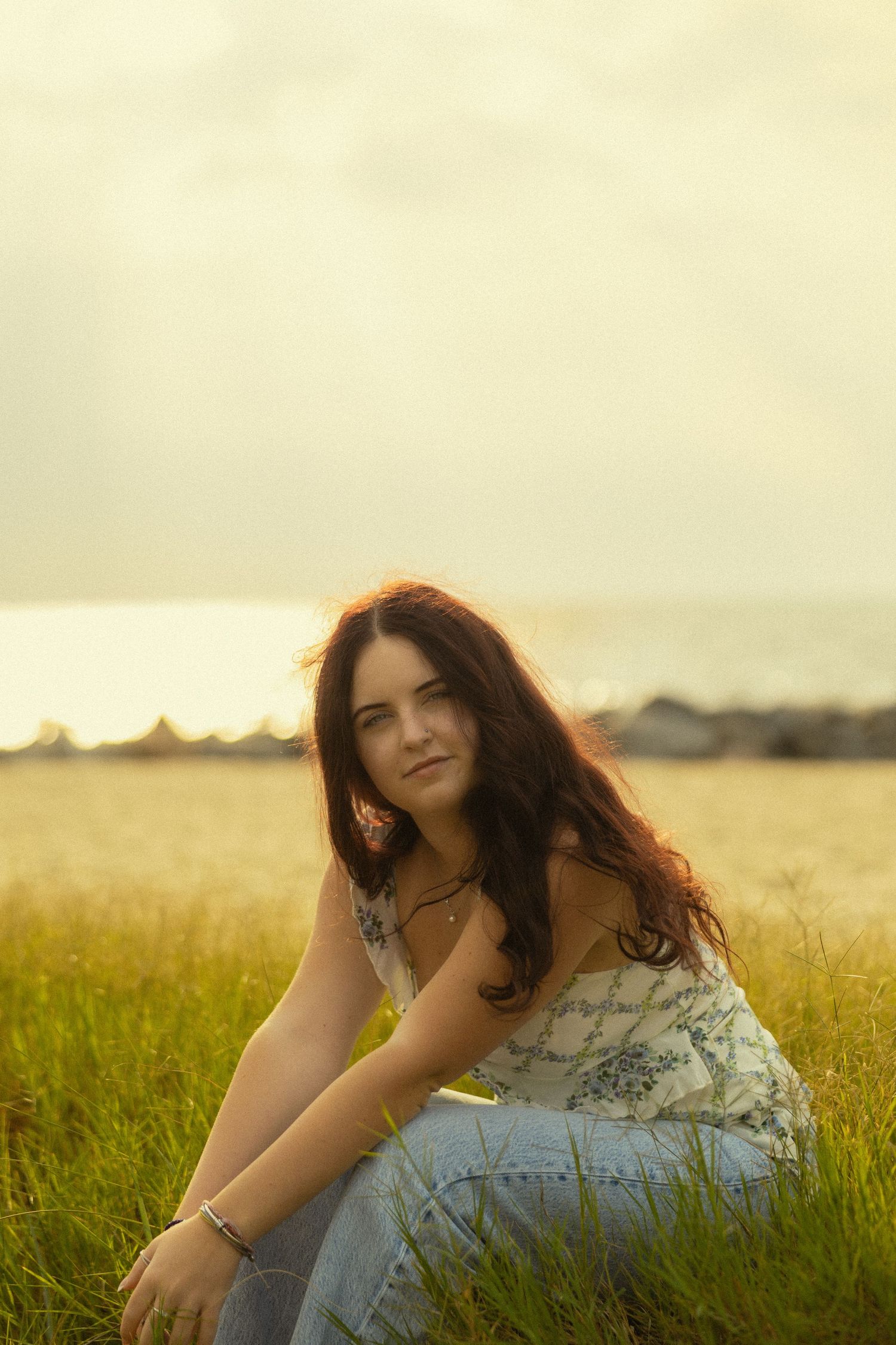 Young woman with long brown hair sitting in golden grass field near ocean at sunset wearing floral dress.