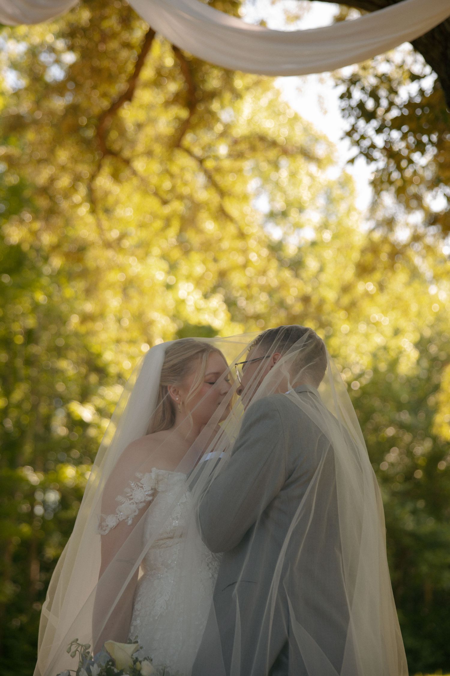 Bride and groom sharing first kiss under floral arch, draped in wedding veil at Avonlea Farms in Mineral, VA. Virginia Wedding Photographer