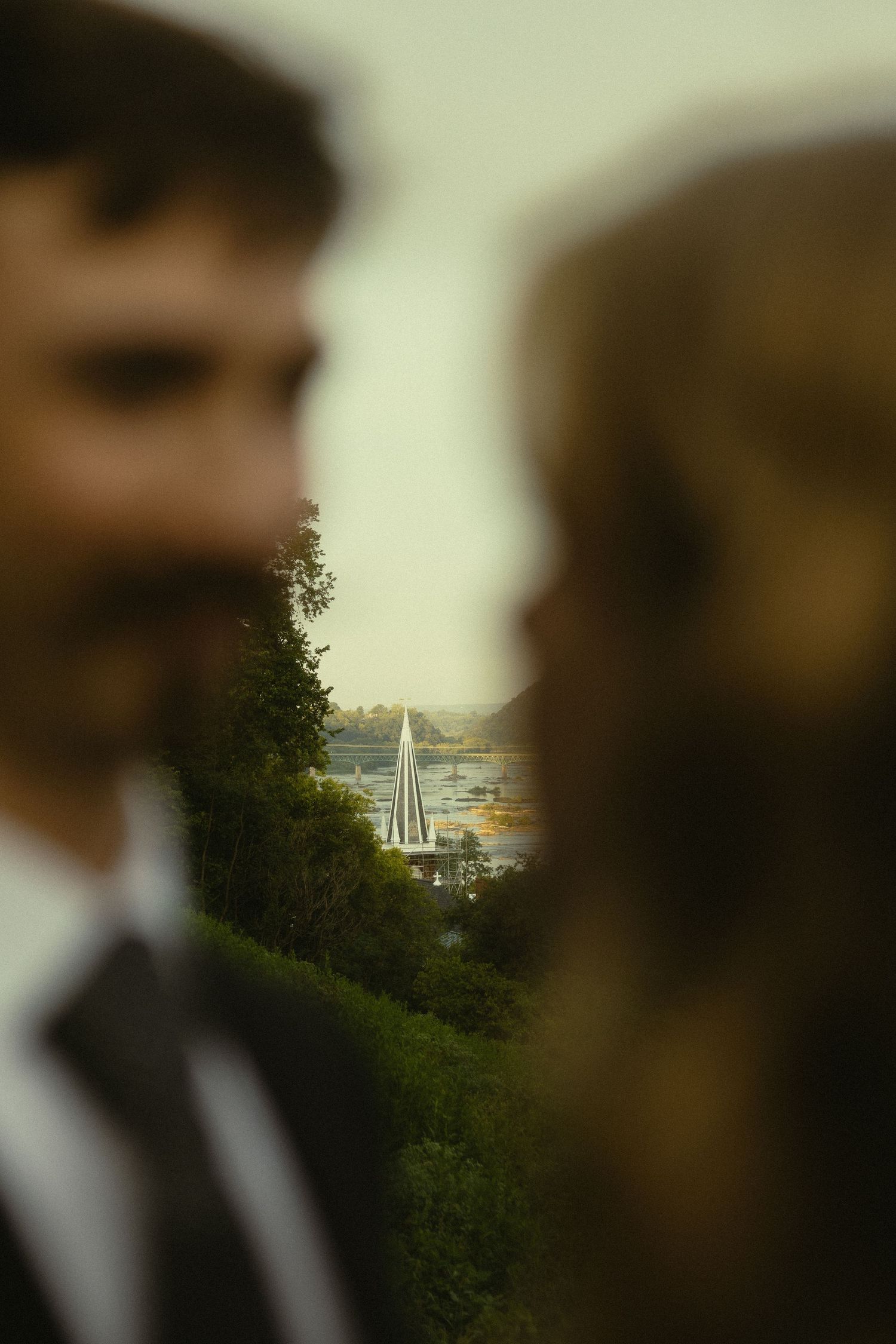 Blurred bride and groom framing a scenic waterfront view with trees and a sailboat in the background.