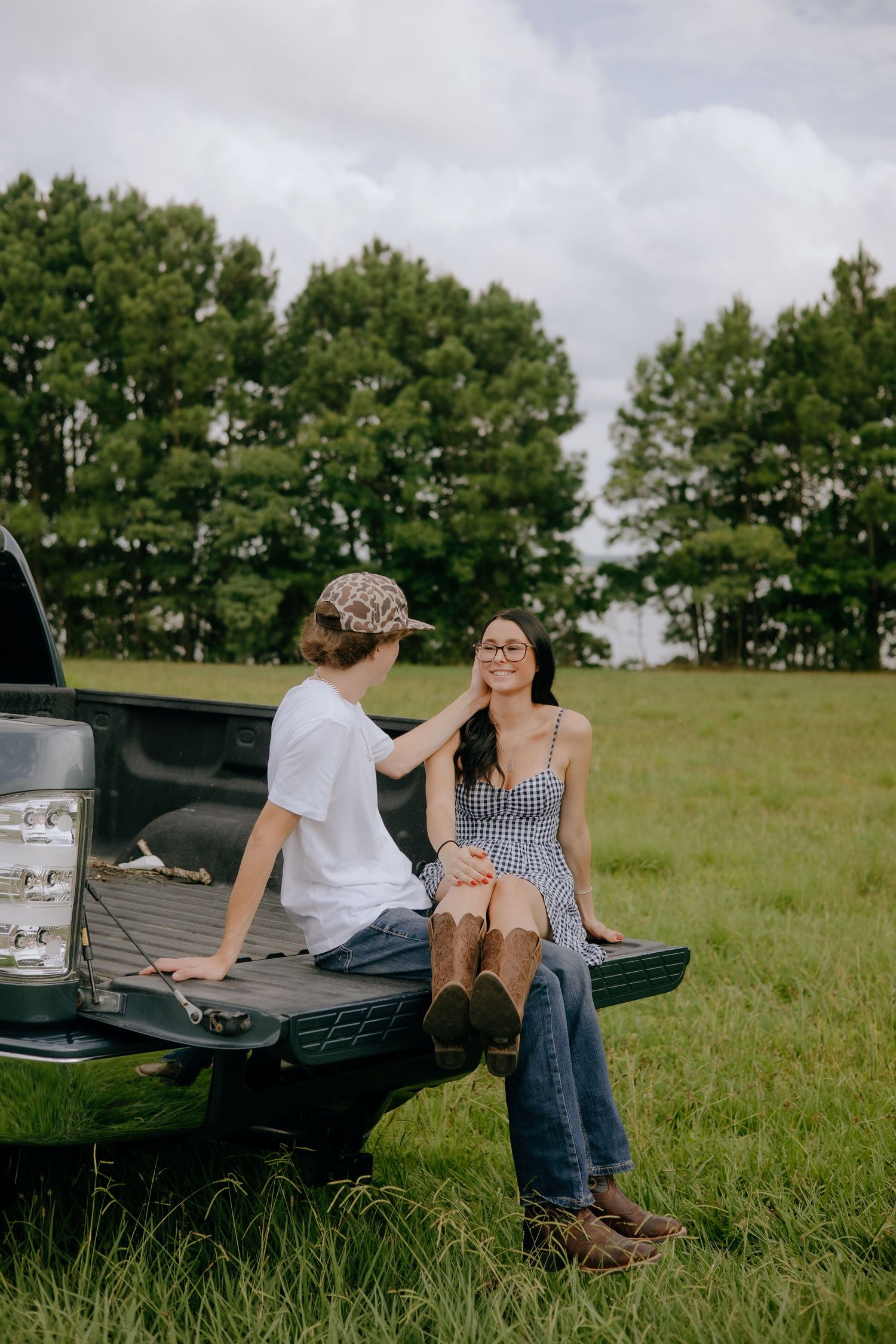 A young couple sits on a truck tailgate in a green field, sharing a tender moment surrounded by trees.