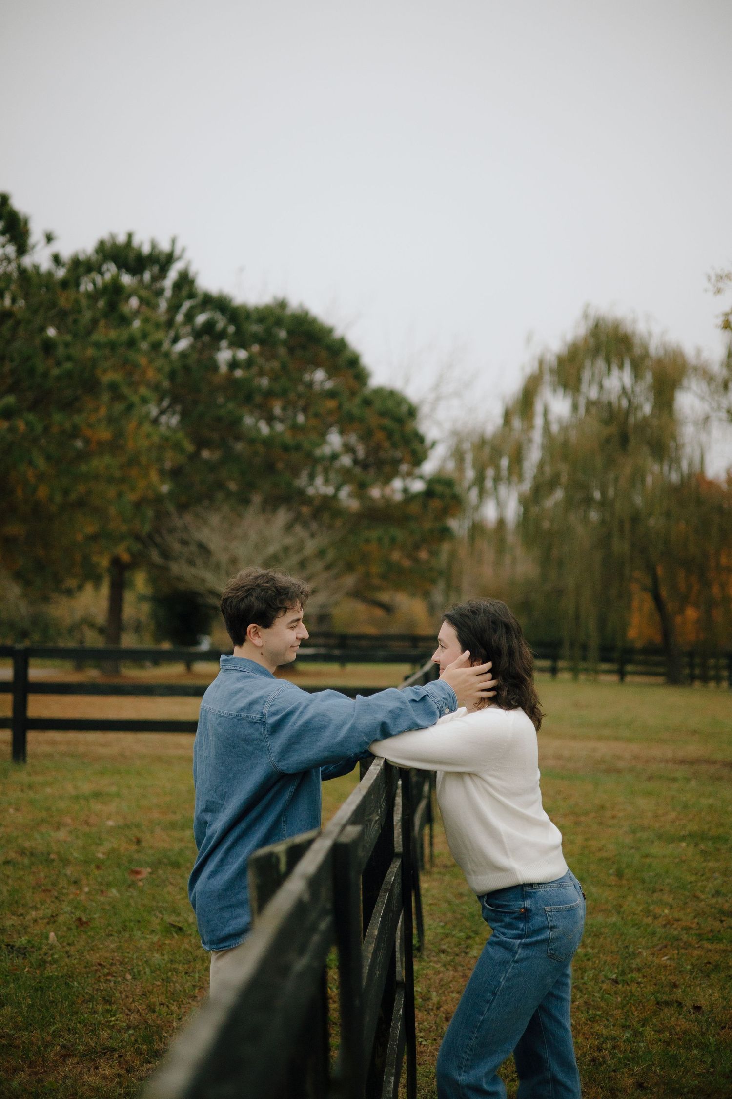 Two people in denim and white clothing playfully interact in a grassy field with autumn trees in the background.