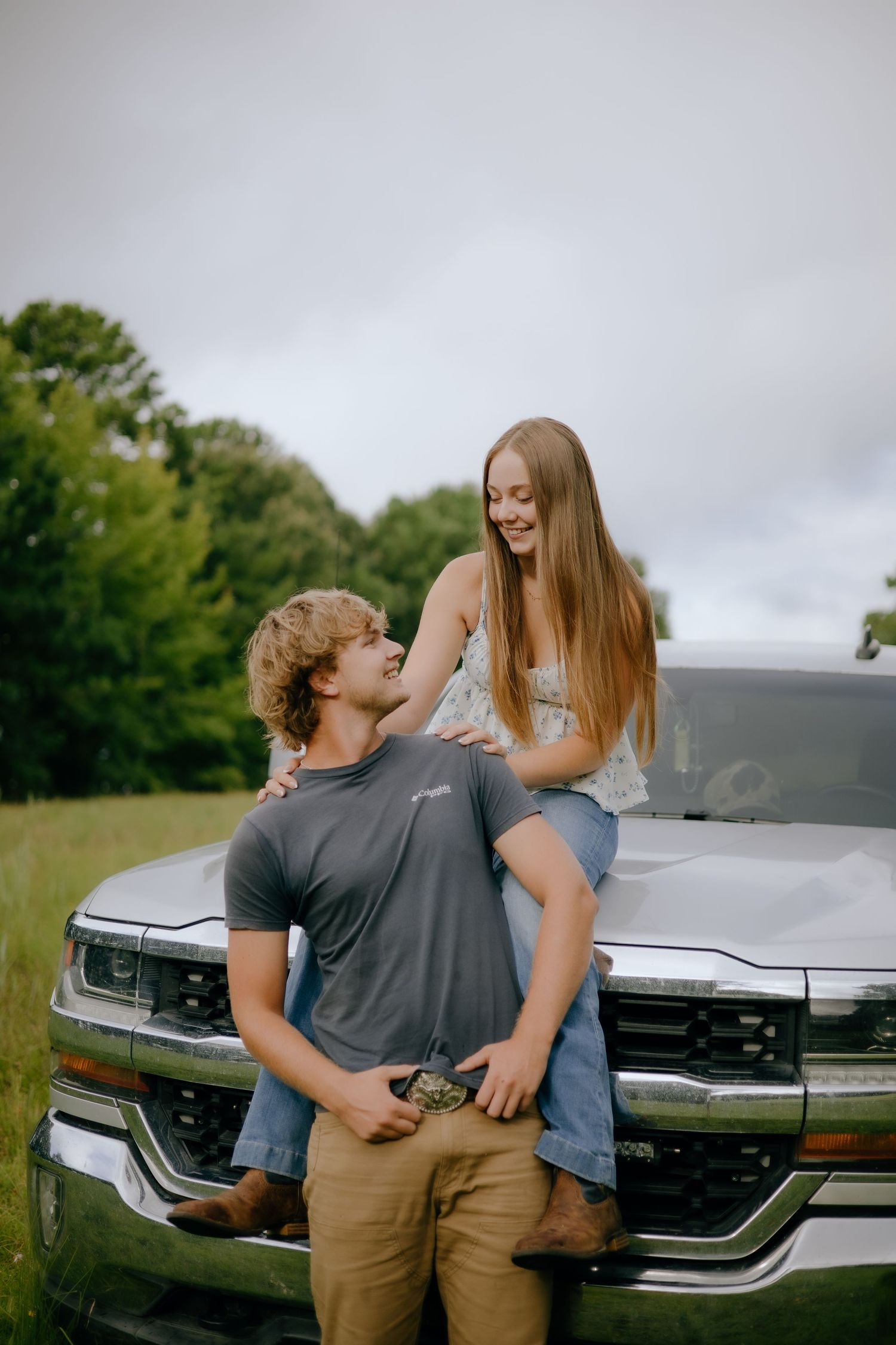 Couple sharing a romantic moment while leaning against a silver pickup truck in a meadow.
