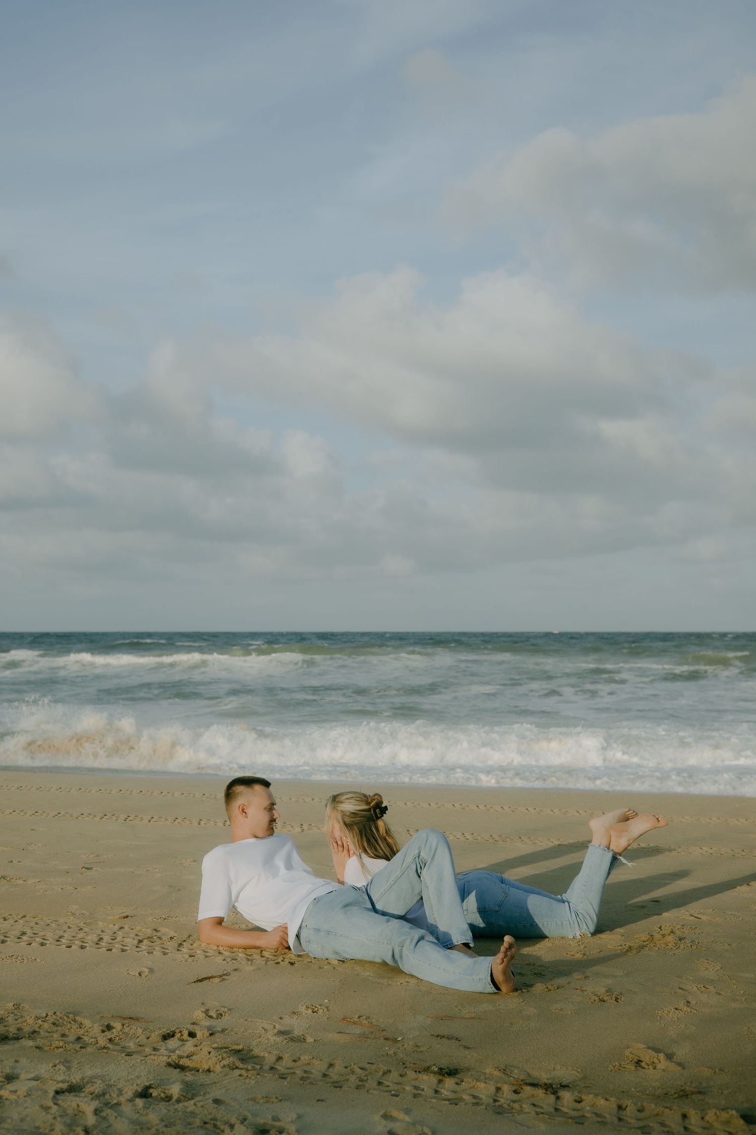 A couple lies relaxed on the sandy beach with ocean waves in the background.