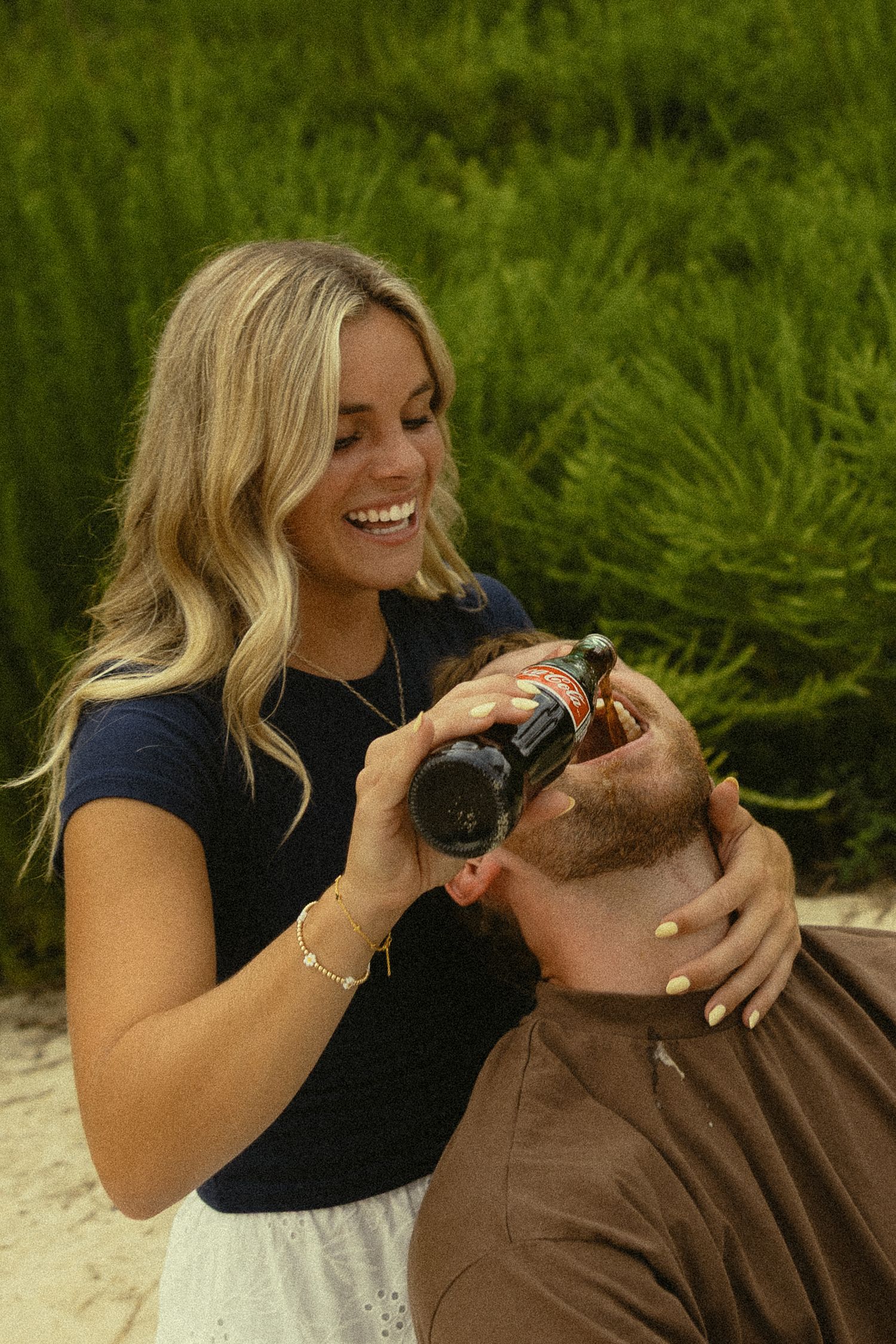 Smiling blonde woman opening a beer bottle outdoors with green bushes in the background.