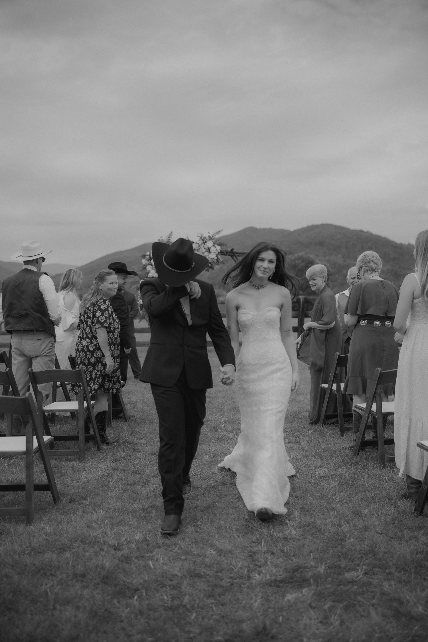 Black and white photo of bride and groom walking down outdoor aisle after wedding ceremony with mountain backdrop.