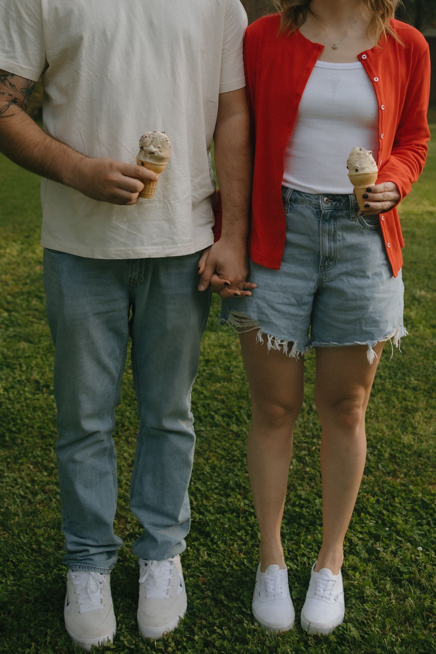 Couple holding hands outdoors on grass, each holding an ice cream cone, wearing jeans and white sneakers.