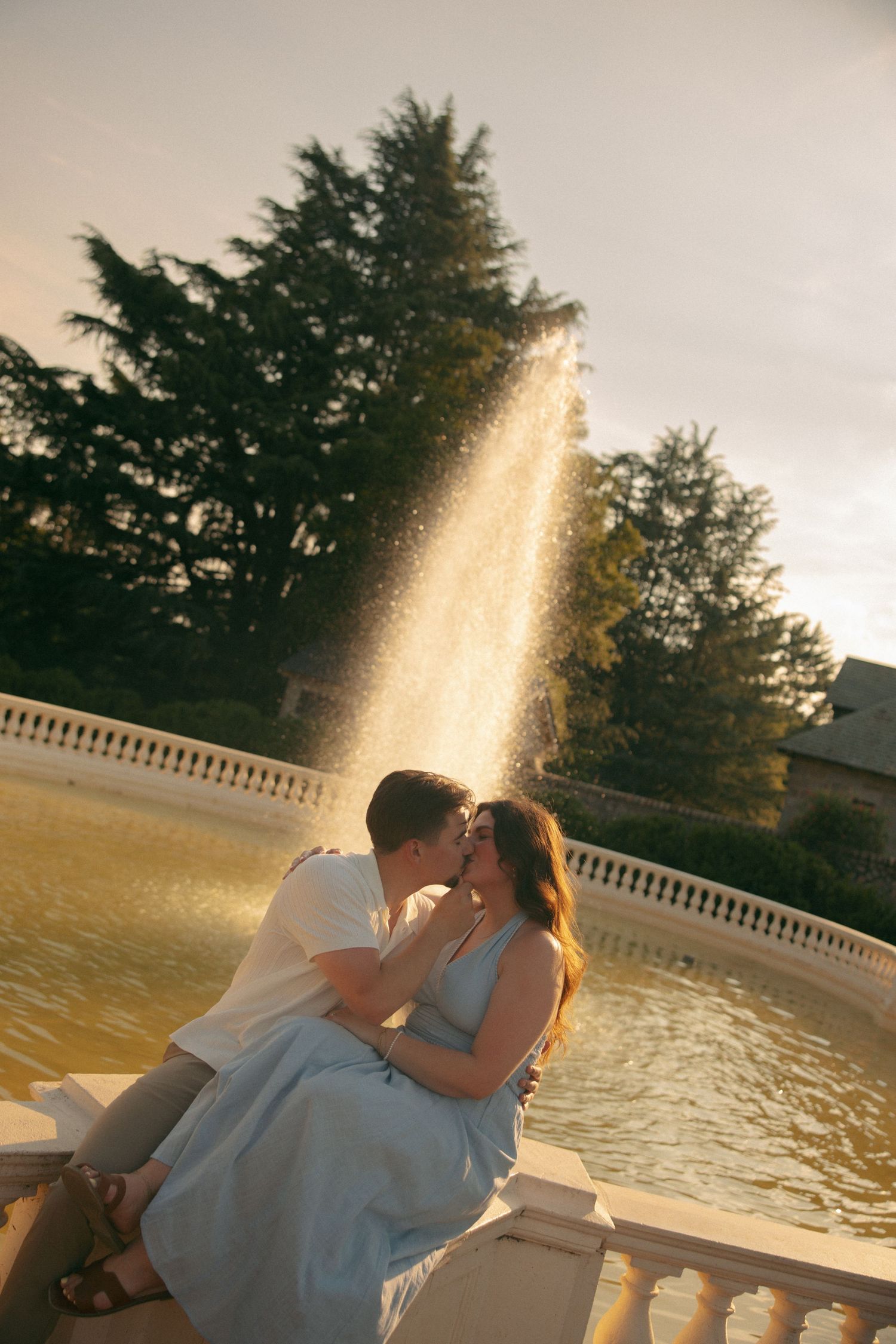 Couple sharing a romantic kiss by a sunlit fountain with golden water and lush trees in the background.