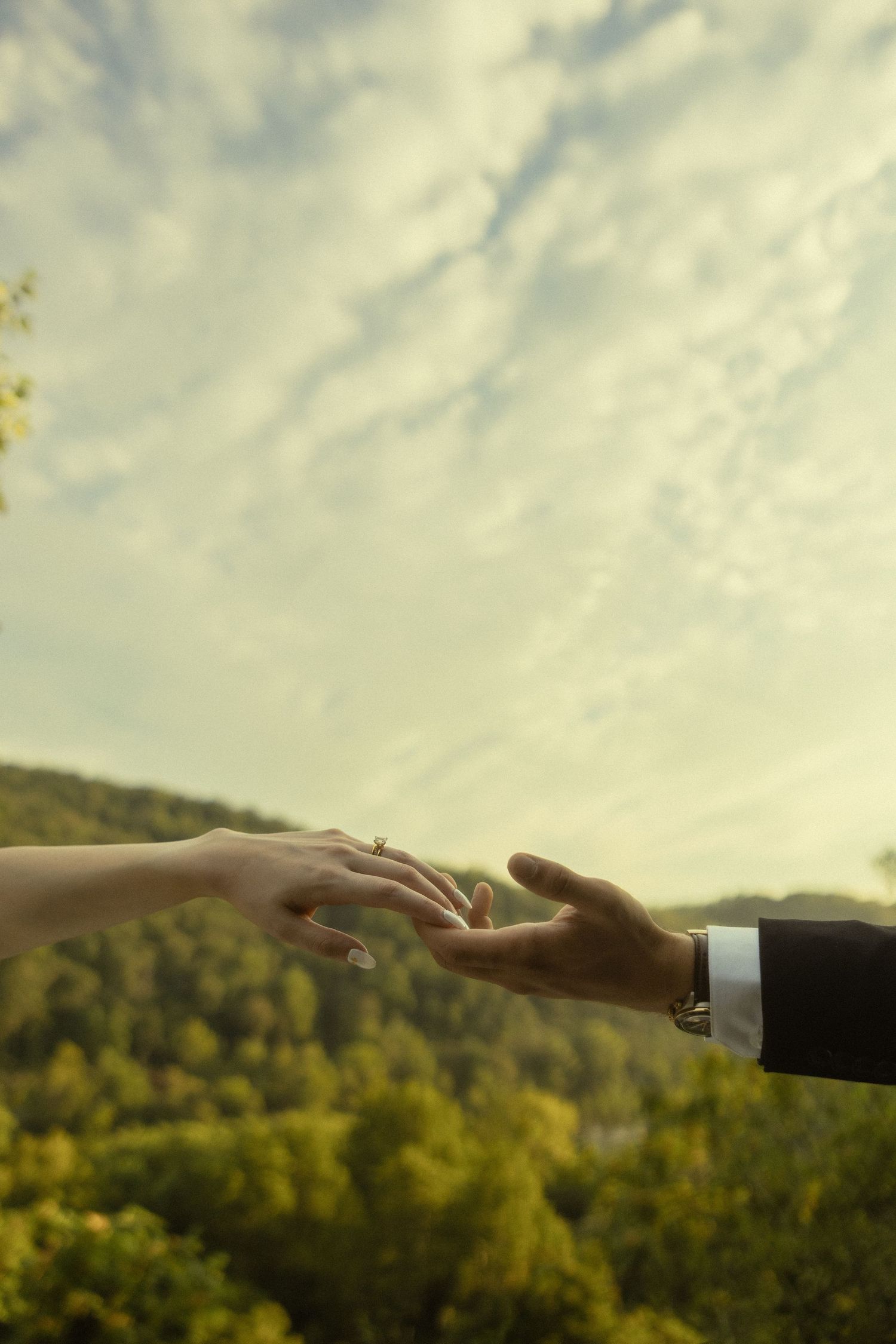 Two hands reaching toward each other against a scenic mountain backdrop, with an engagement ring visible on the woman's finger.