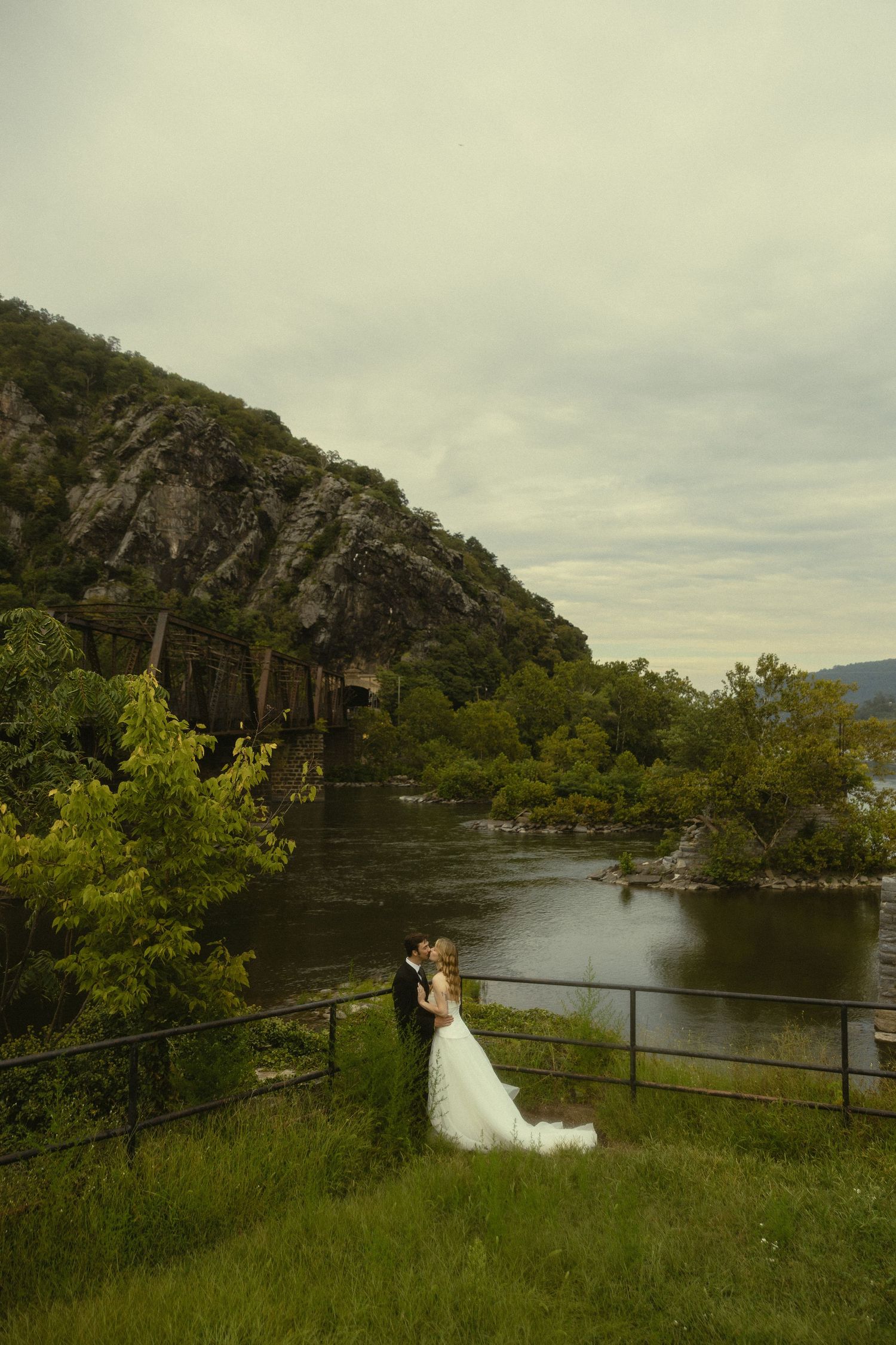 Bride and groom share a romantic kiss overlooking a scenic river and mountain landscape on their wedding day.