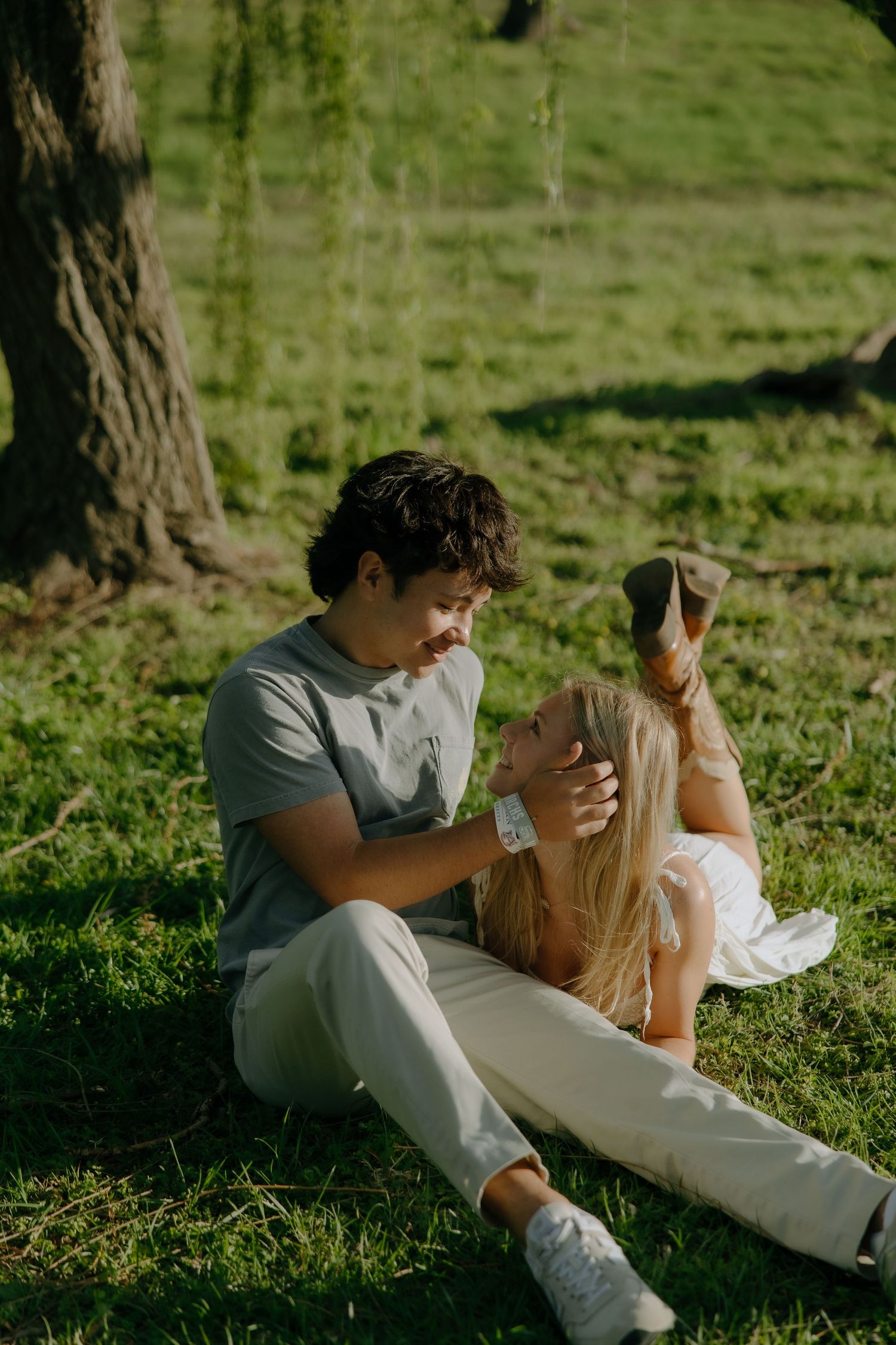 Couple sitting on green grass under a tree, woman resting her head on man's lap in a sunny park.