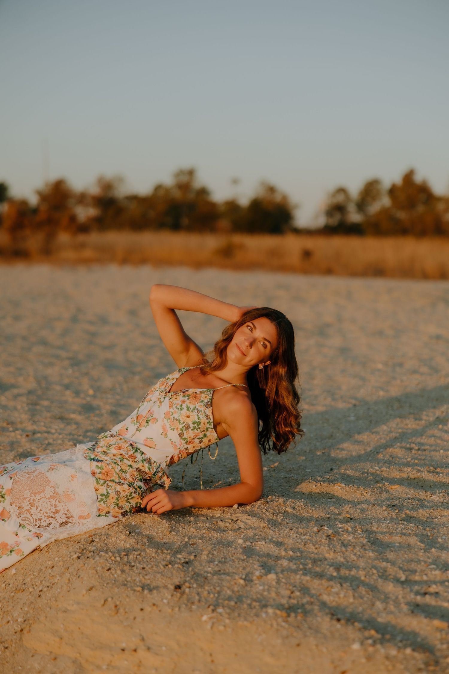 Woman in floral dress lounging on sandy beach at golden hour sunset with trees in background.