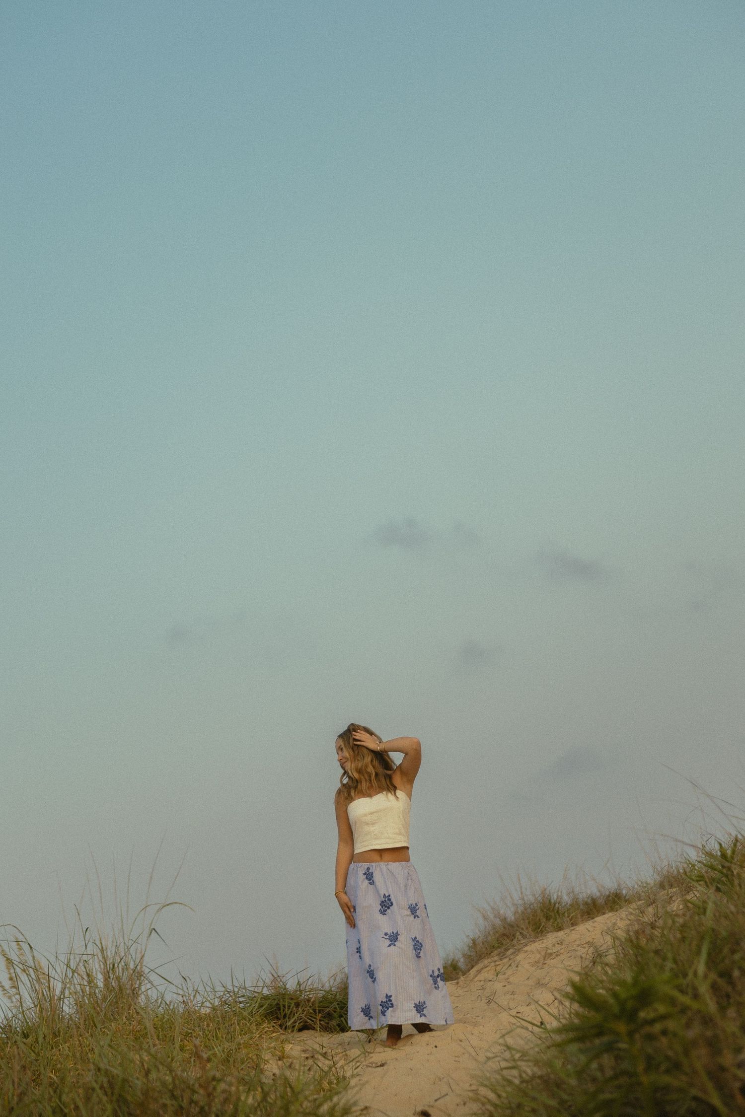 Woman in white crop top and floral maxi skirt standing on sandy dune against blue sky.