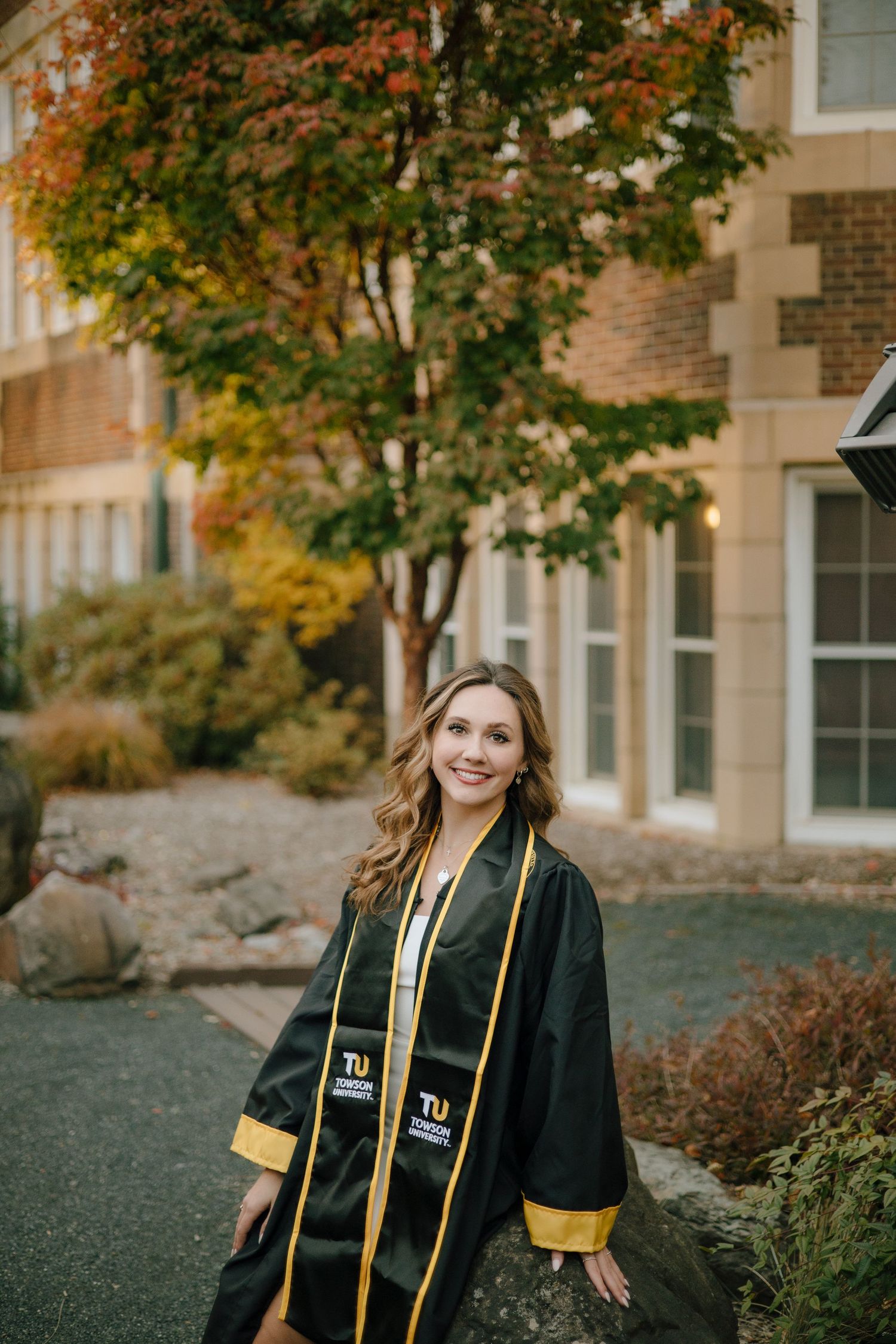 Graduate in black and gold regalia poses outdoors on campus during autumn with colorful fall foliage.