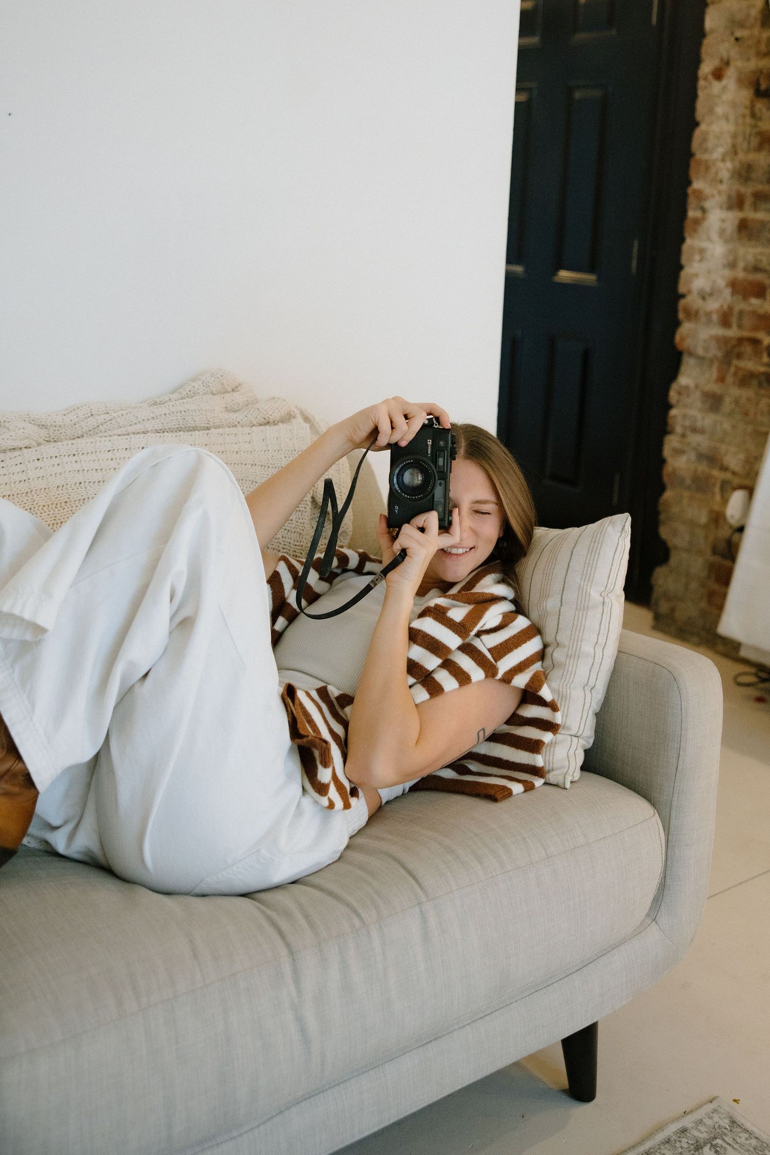 A person in a striped shirt lounges on a light gray couch while taking photos with a vintage camera.